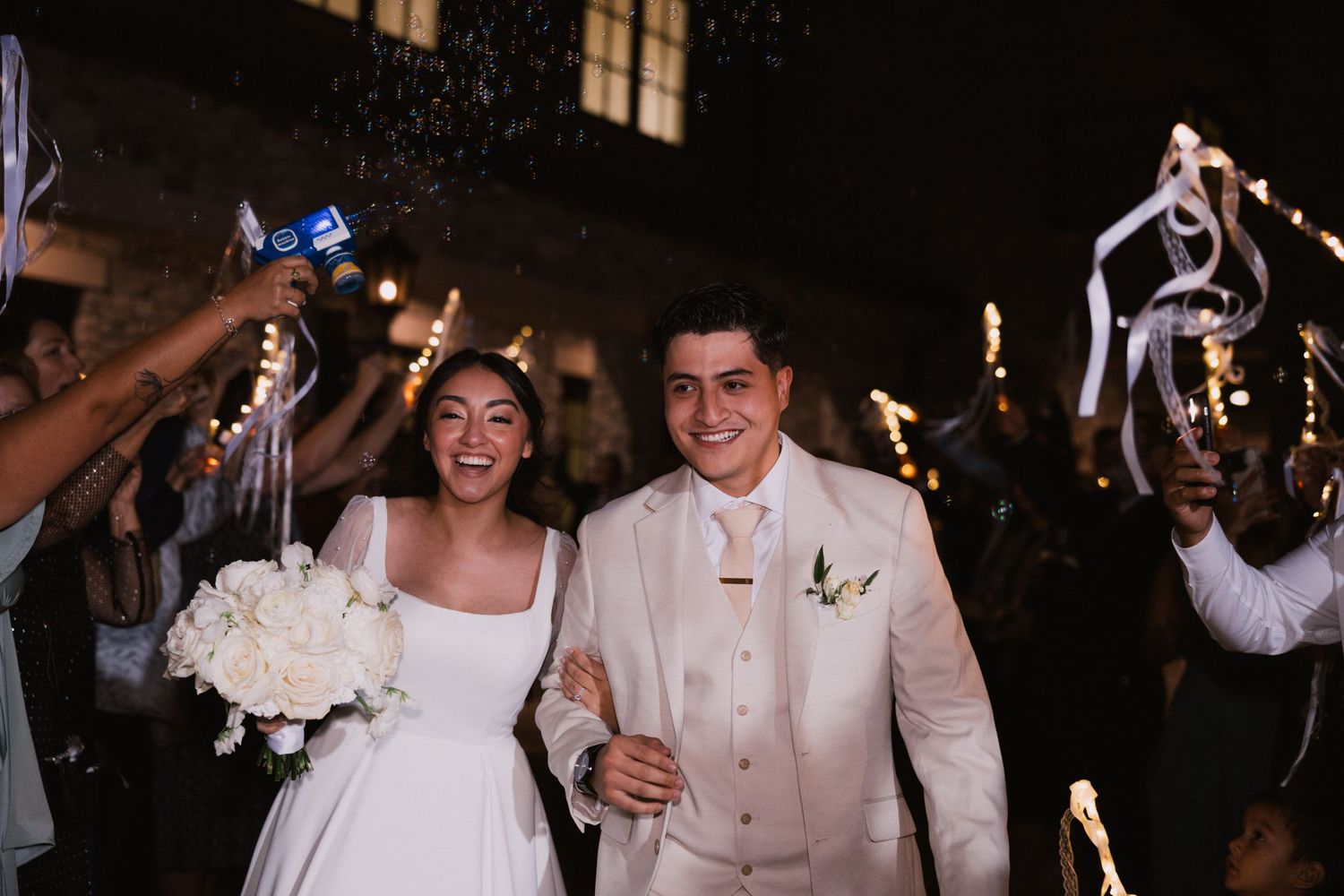 Newlywed couple exits wedding ceremony under sparklers, bride holding white bouquet in elegant gown, groom in white suit.