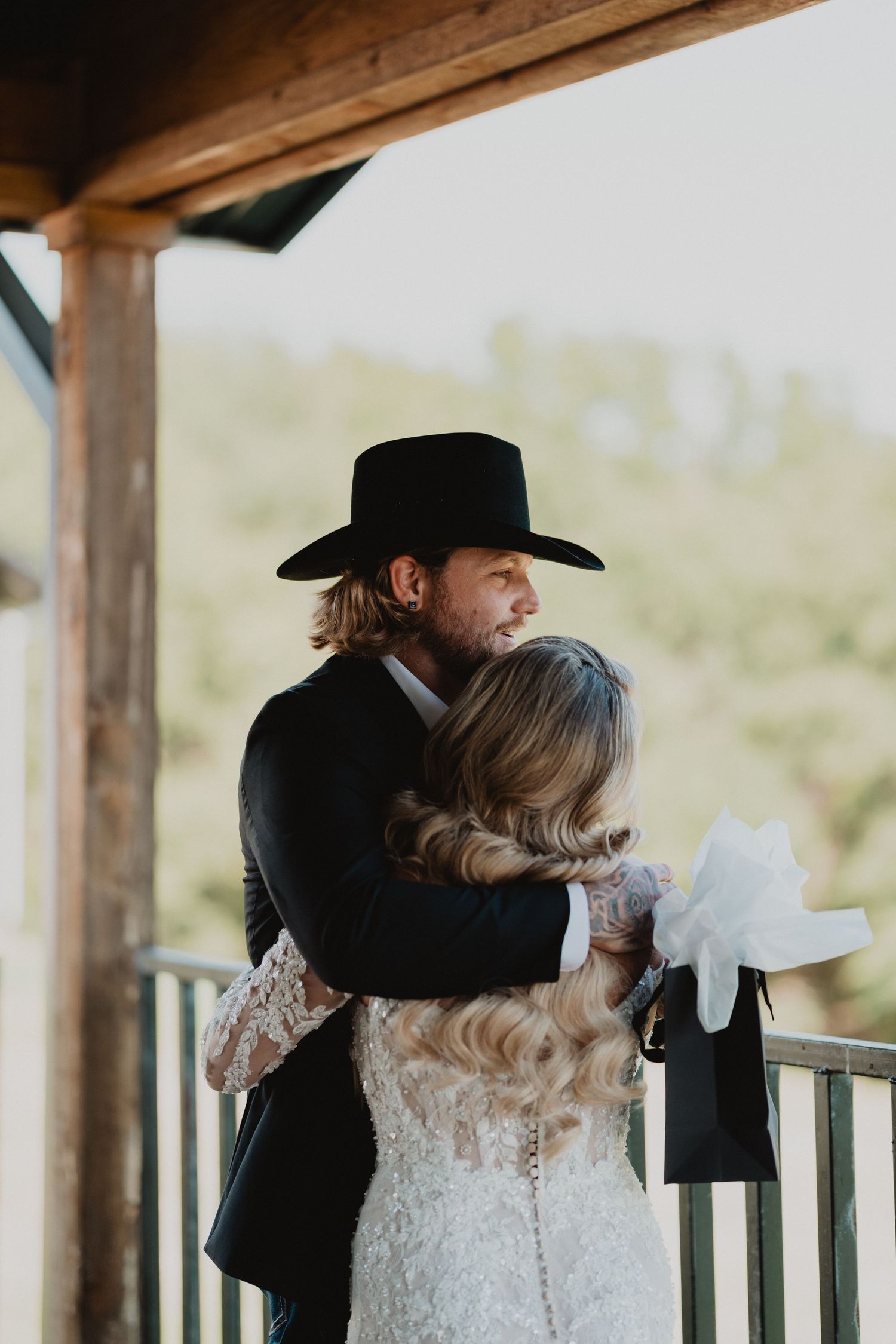 A bride and groom share a tender embrace on a rustic porch, with the groom wearing a black cowboy hat.