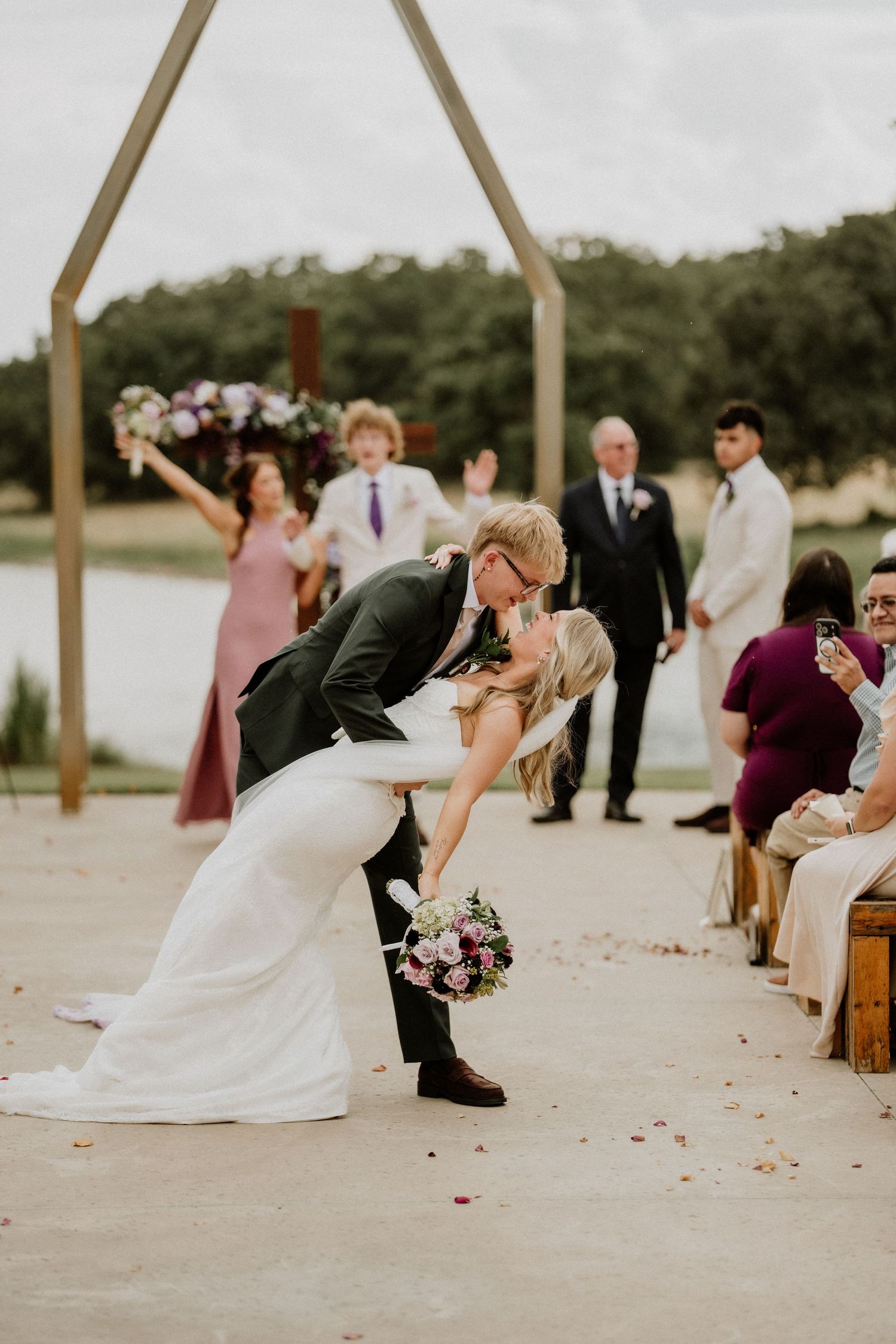 Groom dips bride for a kiss at outdoor wedding ceremony with wooden arch, bridesmaids, and guests cheering.