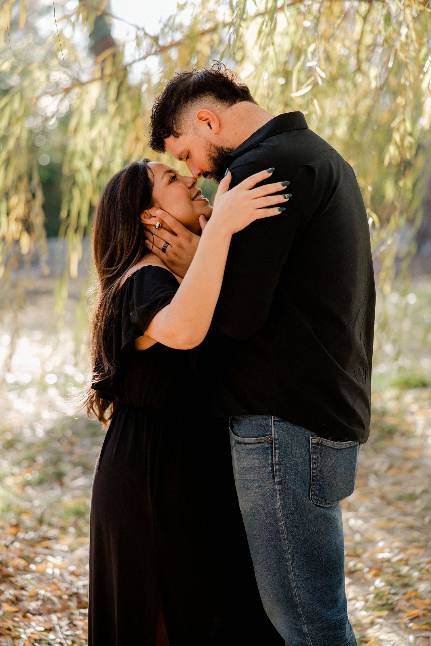 Couple sharing a romantic kiss under weeping willow branches in golden hour light, both dressed in black.