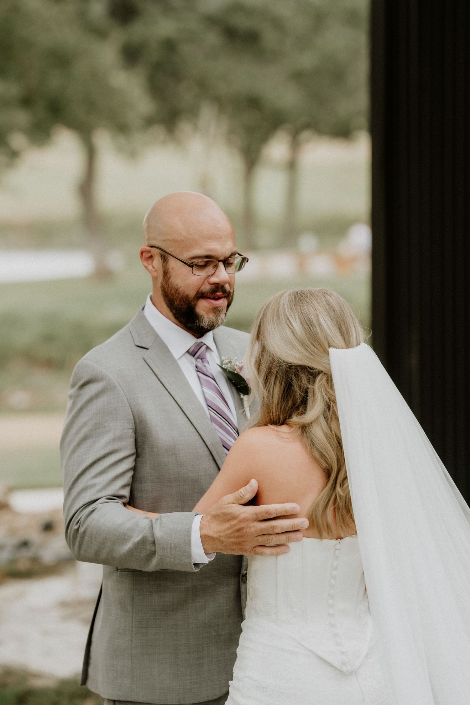 Groom in gray suit smiling at bride in white dress and veil during wedding first look outdoors.