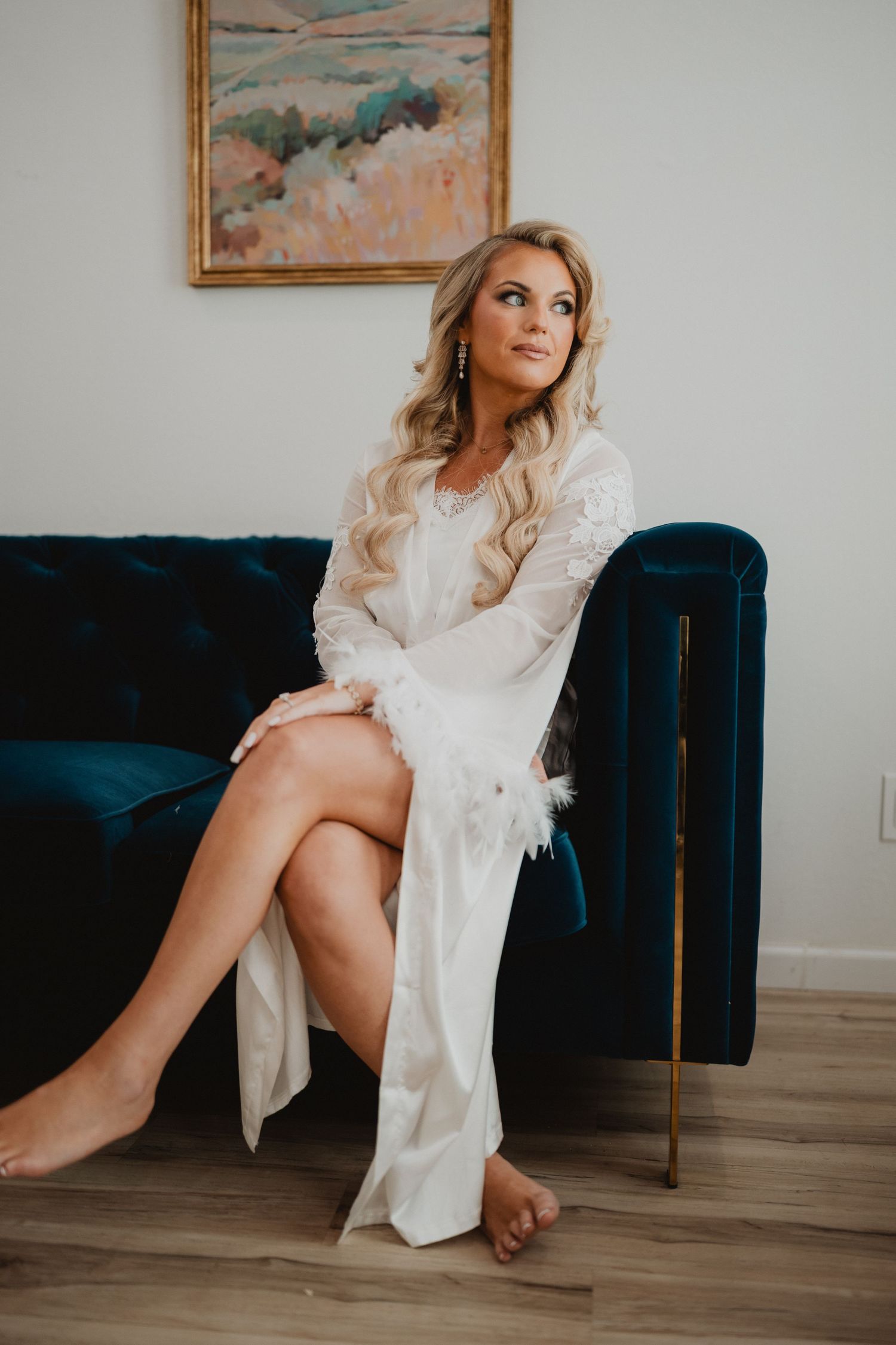 Blonde bride in white lace robe sits elegantly on a teal velvet sofa, gazing upward in a bright, modern room.