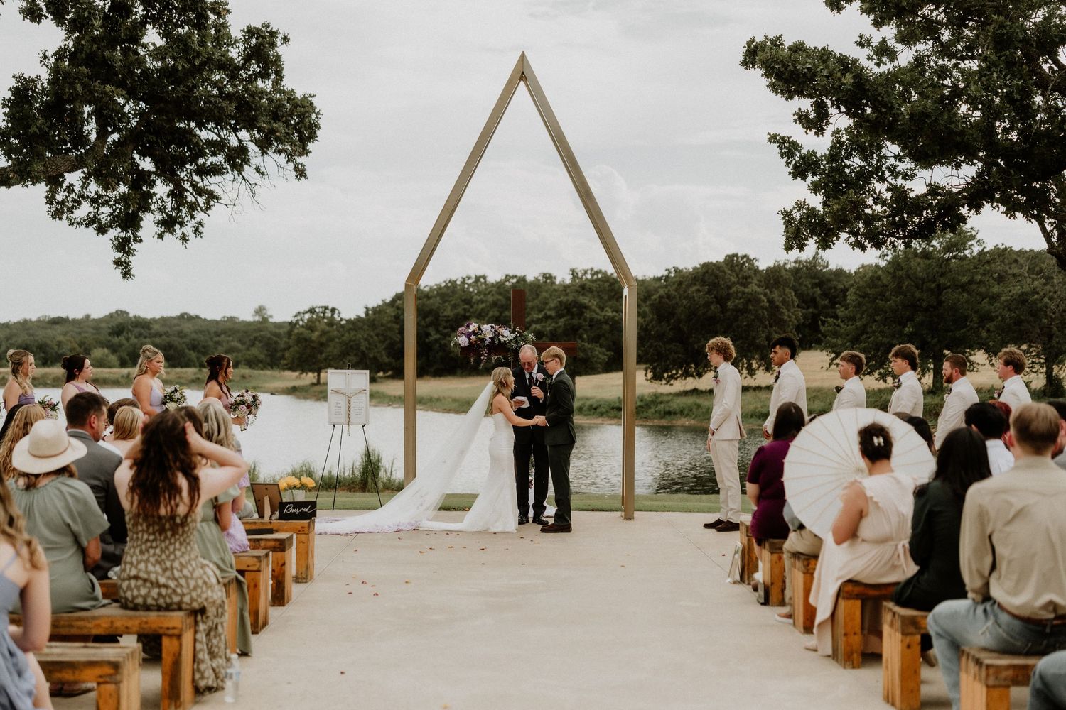 Outdoor wedding ceremony with triangular wooden arch, bride and groom, guests seated on wooden benches near a serene lake.