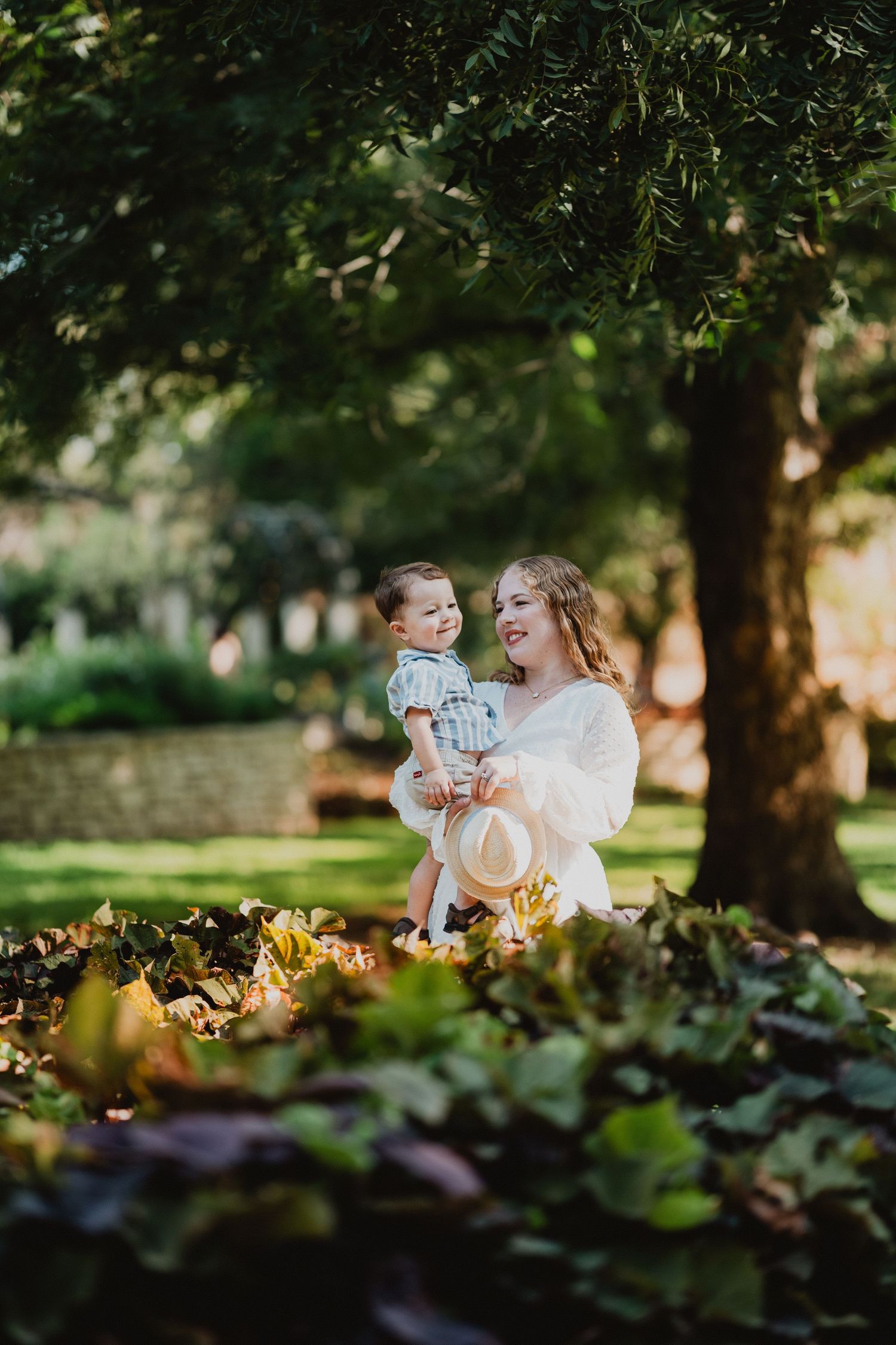 Mother in white dress holding toddler under a large tree in a green garden, both smiling warmly.