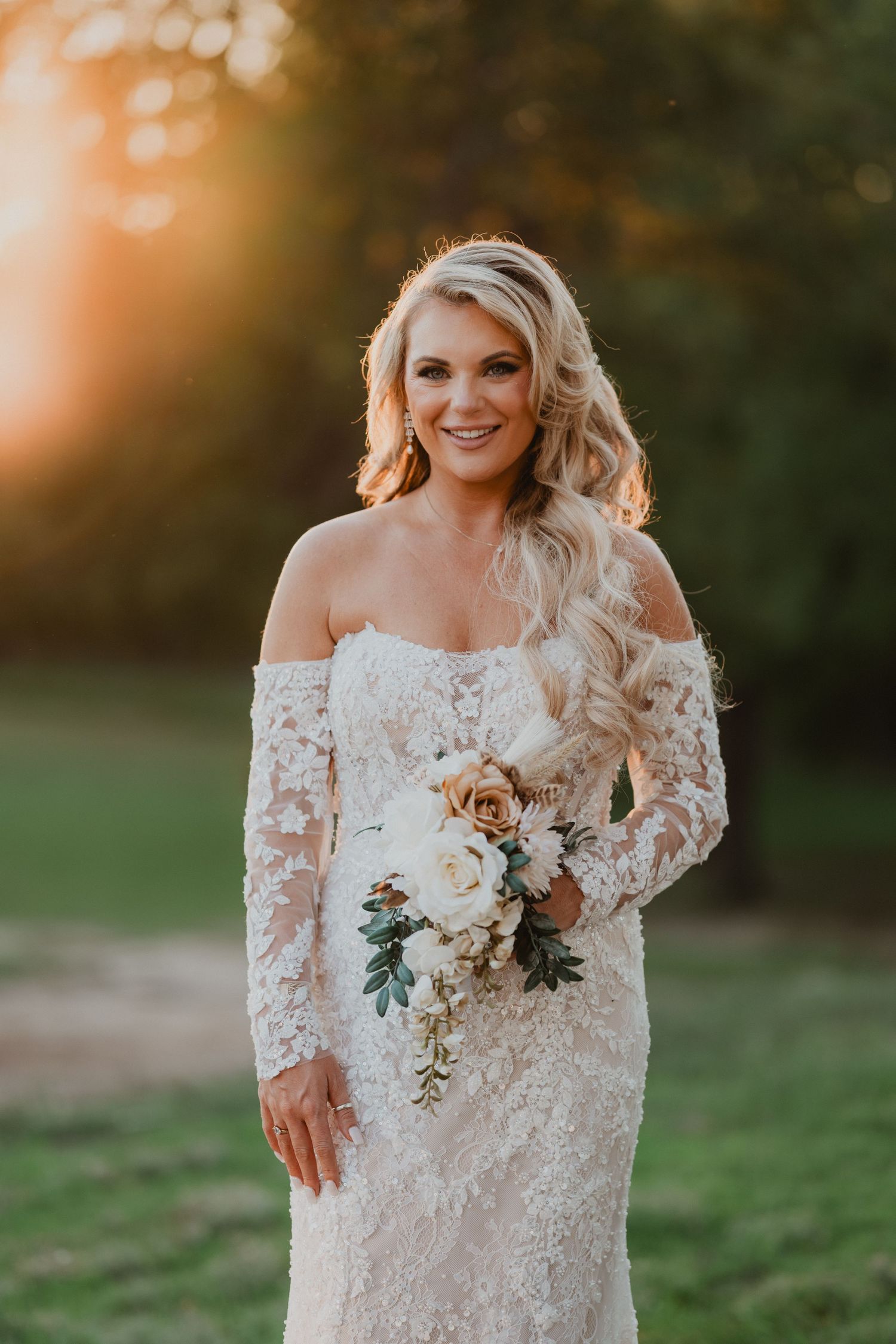 Bride in lace off-shoulder gown holding white floral bouquet, golden sunset backdrop.