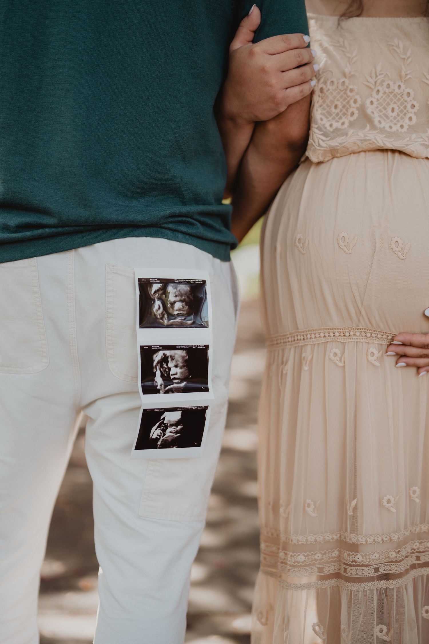 Close-up of couple holding baby bump with ultrasound photos tucked in man's white pants pocket.