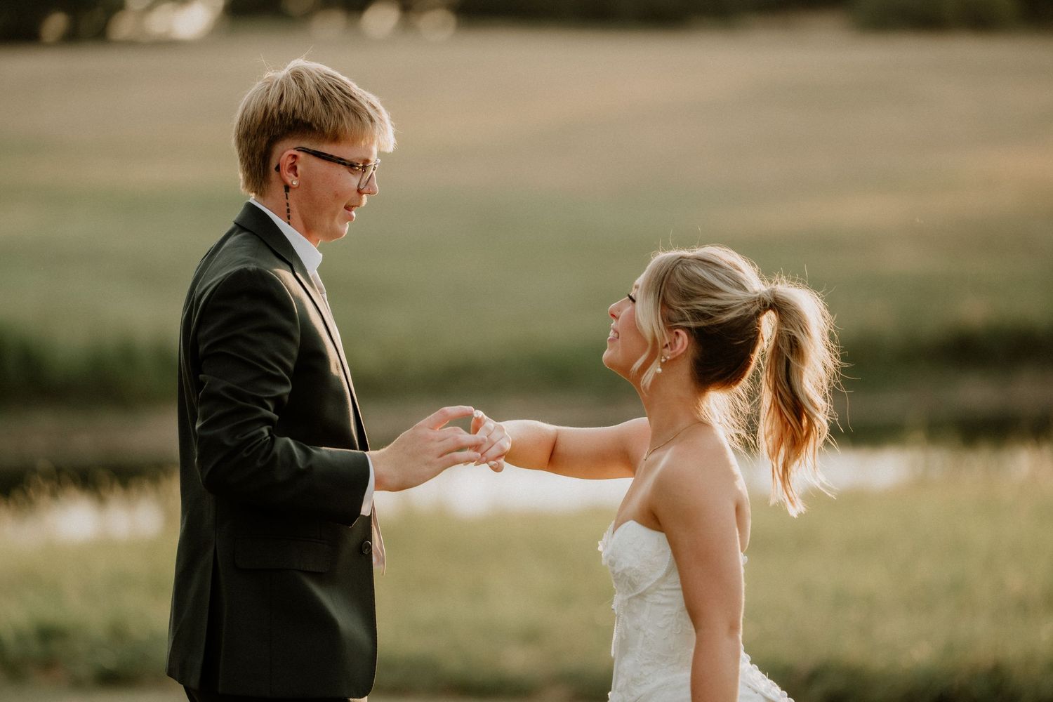 Bride in white strapless gown holding hands with groom in dark suit at golden hour in a green field.