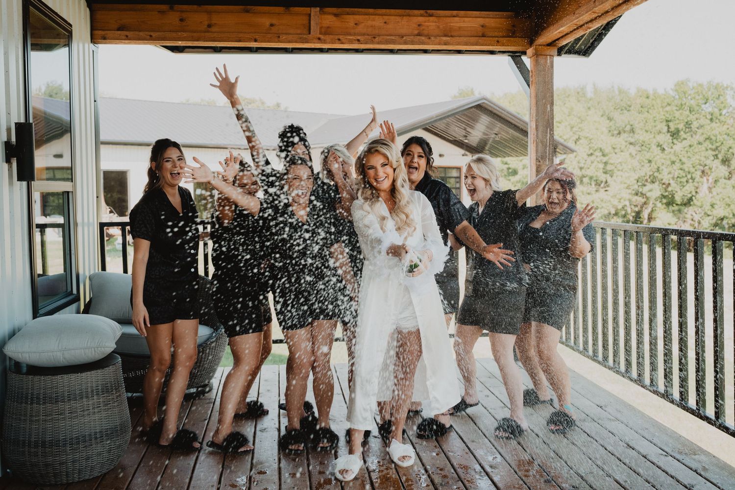 Bride in white robe celebrates with bridesmaids in black robes tossing confetti on a wooden deck.