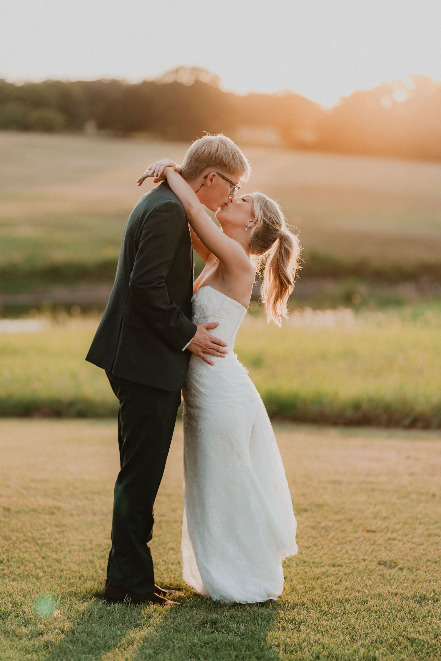 Bride and groom sharing a romantic sunset kiss on green grass field, bride in white gown, groom in dark suit.