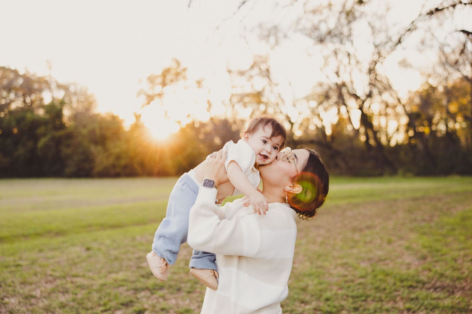 Mother lifts laughing baby in the air during a golden hour outdoor photo session in a grassy park.