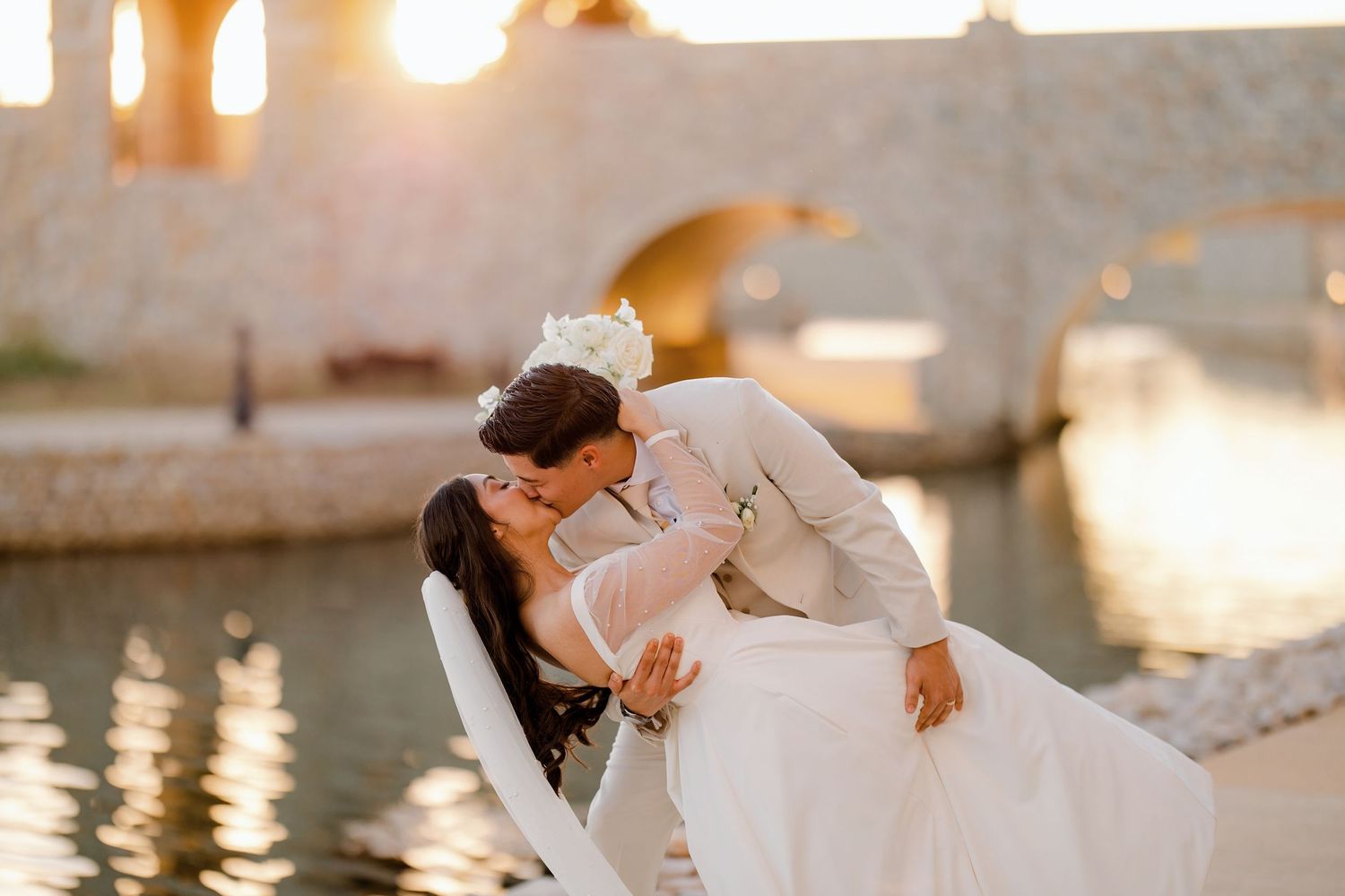 Bride and groom share a passionate dip kiss at sunset near a beautiful stone arch bridge.