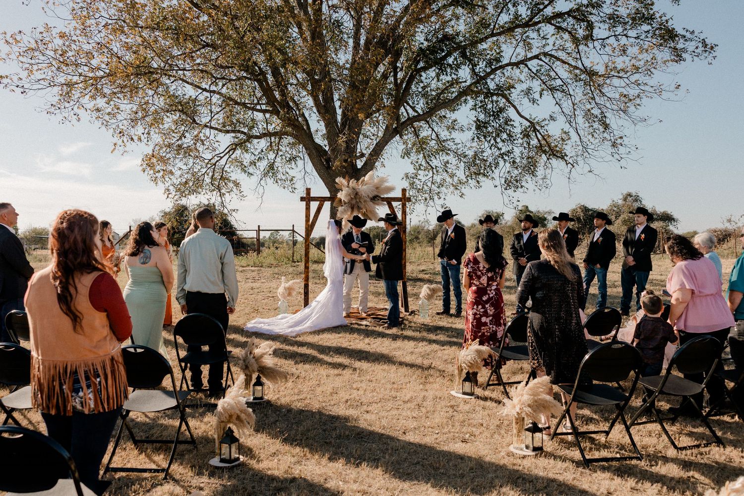 Outdoor wedding ceremony under a large tree with guests seated on chairs in a rustic countryside setting.