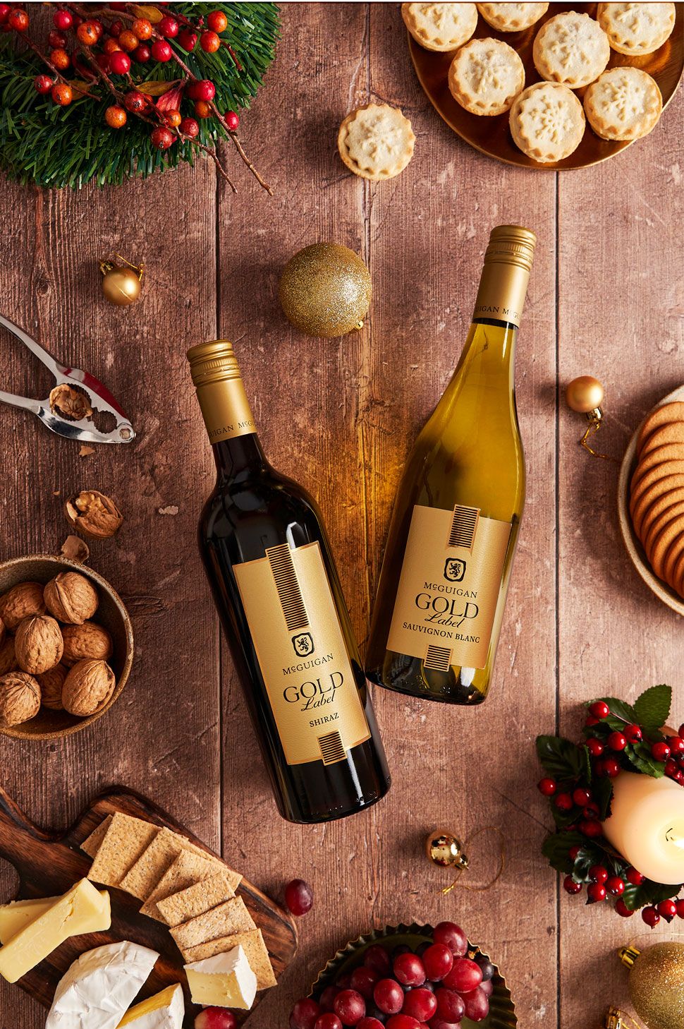 Two gold-labeled wine bottles, Shiraz and Sauvignon Blanc, displayed on a wooden table with festive Christmas decorations.