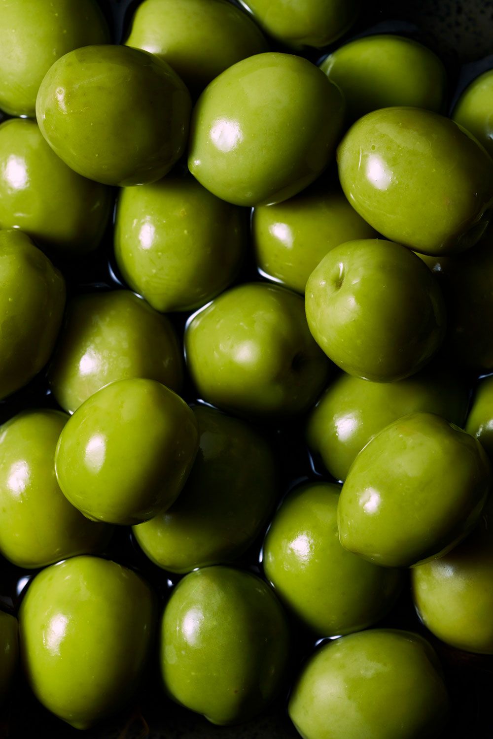 Close-up of fresh, glossy green olives piled together, glistening with brine in natural light.