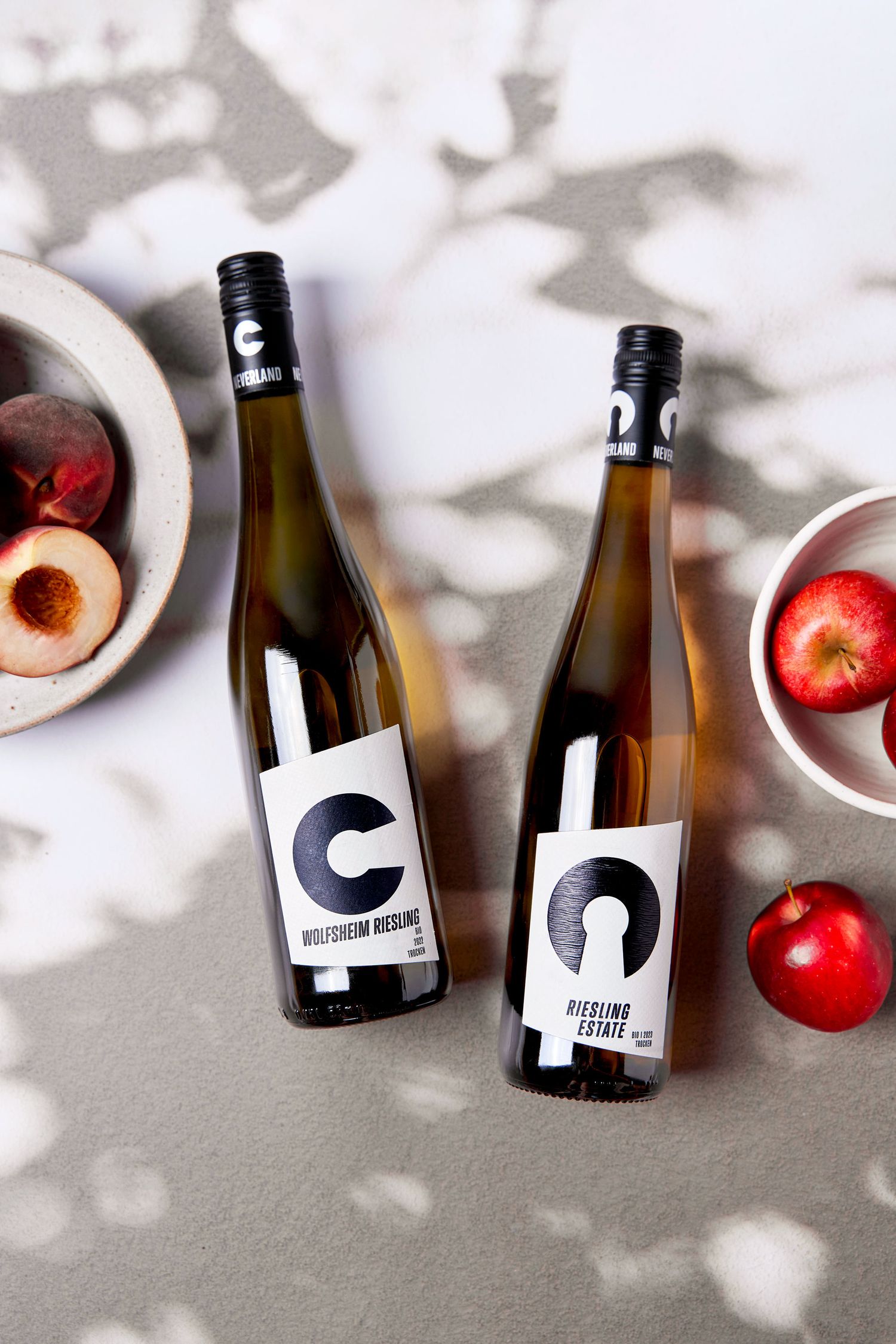 Two bottles of Riesling wine beside bowls of fresh red apples and nectarines on a white shadowed surface.