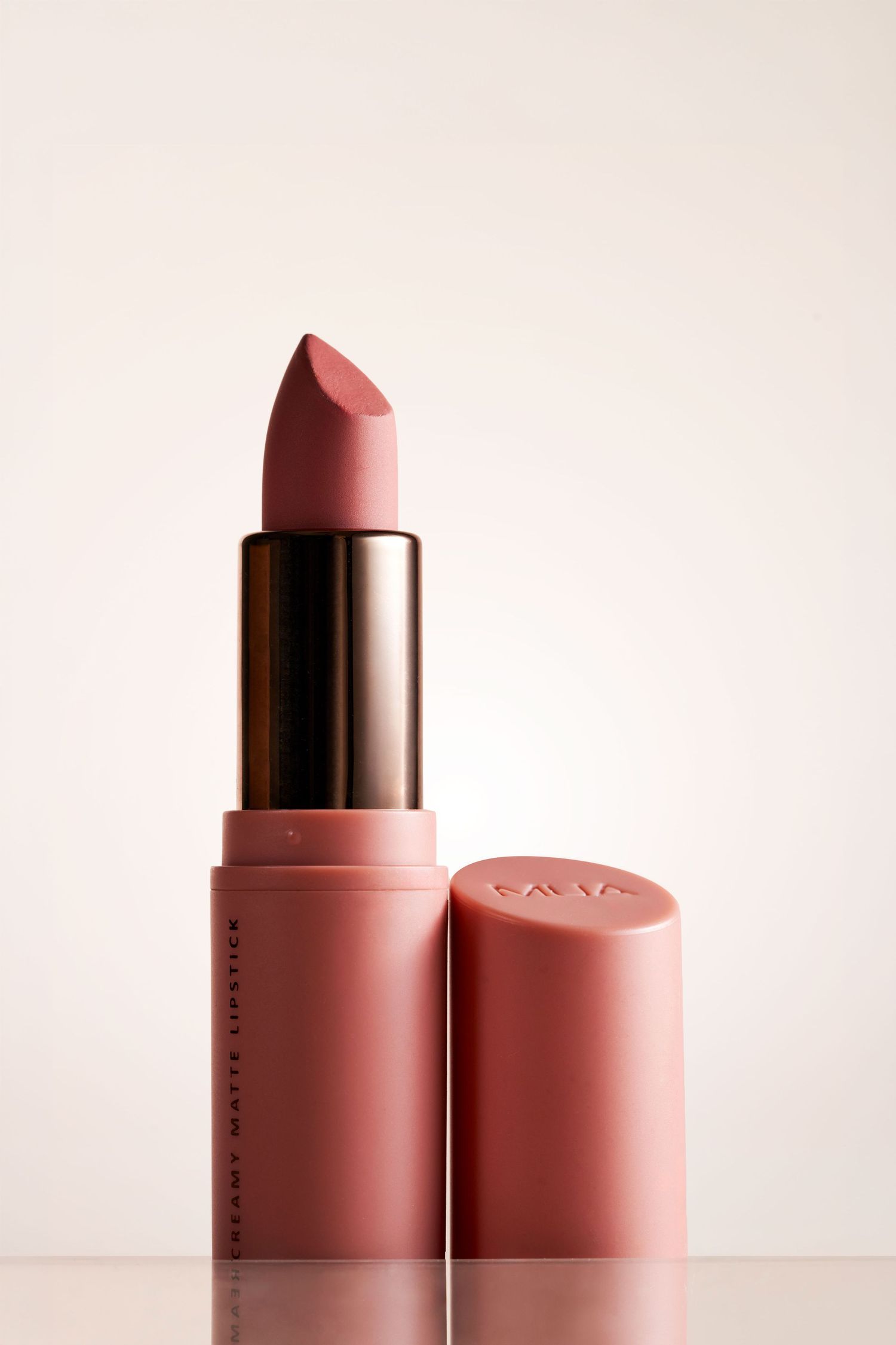 Creamy matte pink lipstick in rose packaging with cap beside it on a light reflective surface.