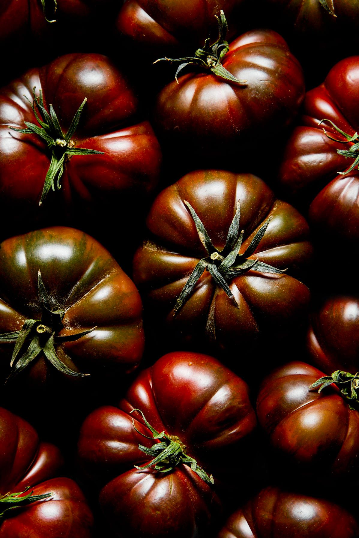 Dark heirloom tomatoes with green stems arranged in a close-up display, showcasing rich burgundy and deep red tones.