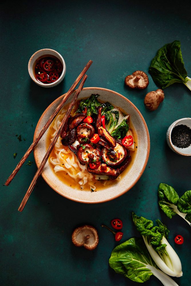 Asian noodle soup bowl with mushrooms, bok choy, chili, and chopsticks on dark teal background.