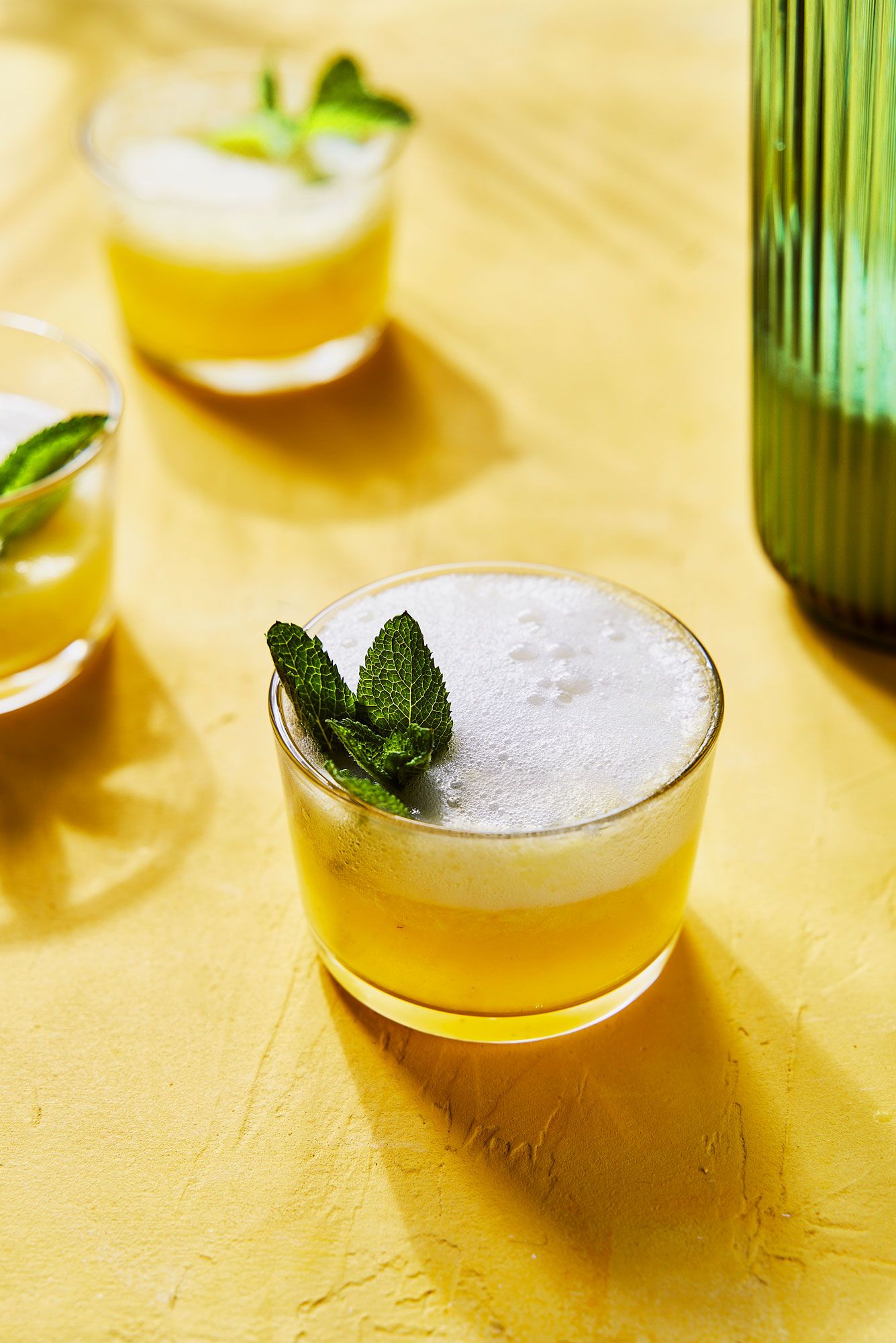 Refreshing yellow cocktails with mint garnish in rocks glasses on a golden surface with a green bottle.