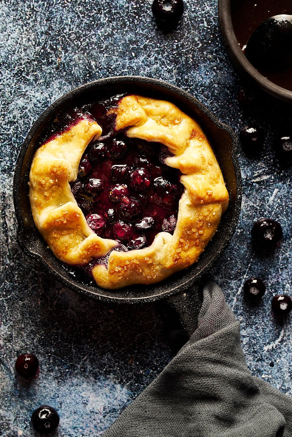 Rustic blueberry galette in cast iron skillet with golden crust, fresh berries scattered on blue surface.