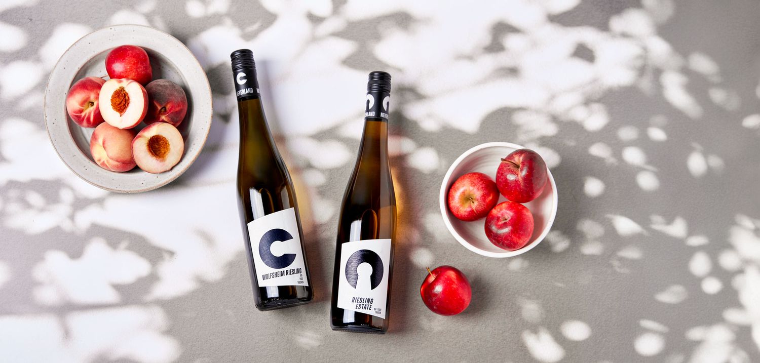 Two wine bottles flanked by bowls of fresh red apples on a light surface with soft dappled shadows.