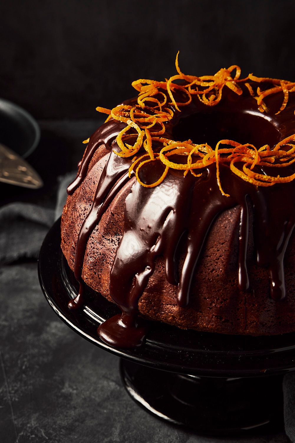 Chocolate bundt cake with dark ganache glaze topped with candied orange zest curls on a black cake stand.