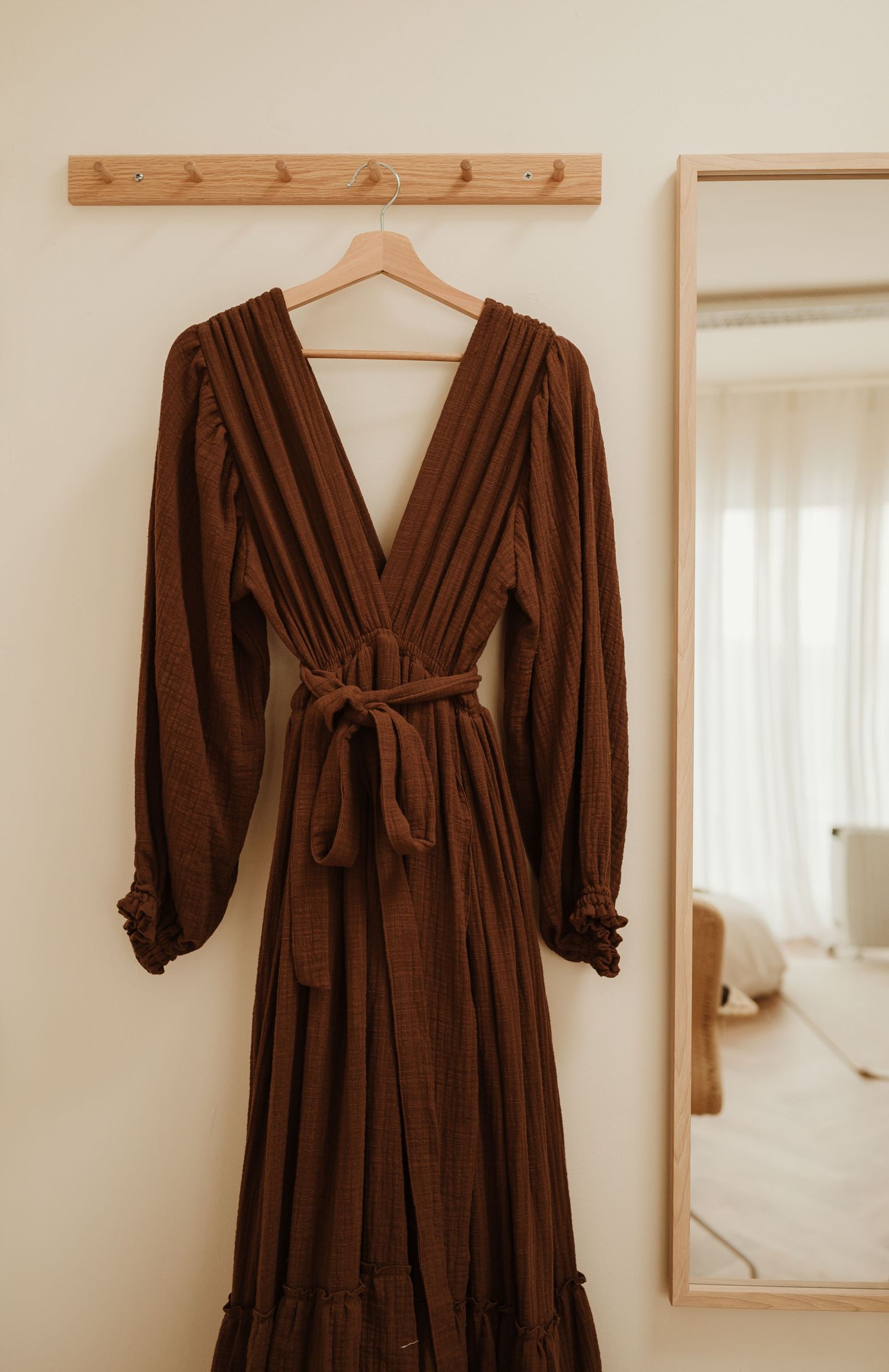 A brown long-sleeve maxi dress with ruffle details and waist tie hanging on a wooden coat hanger against a cream wall.