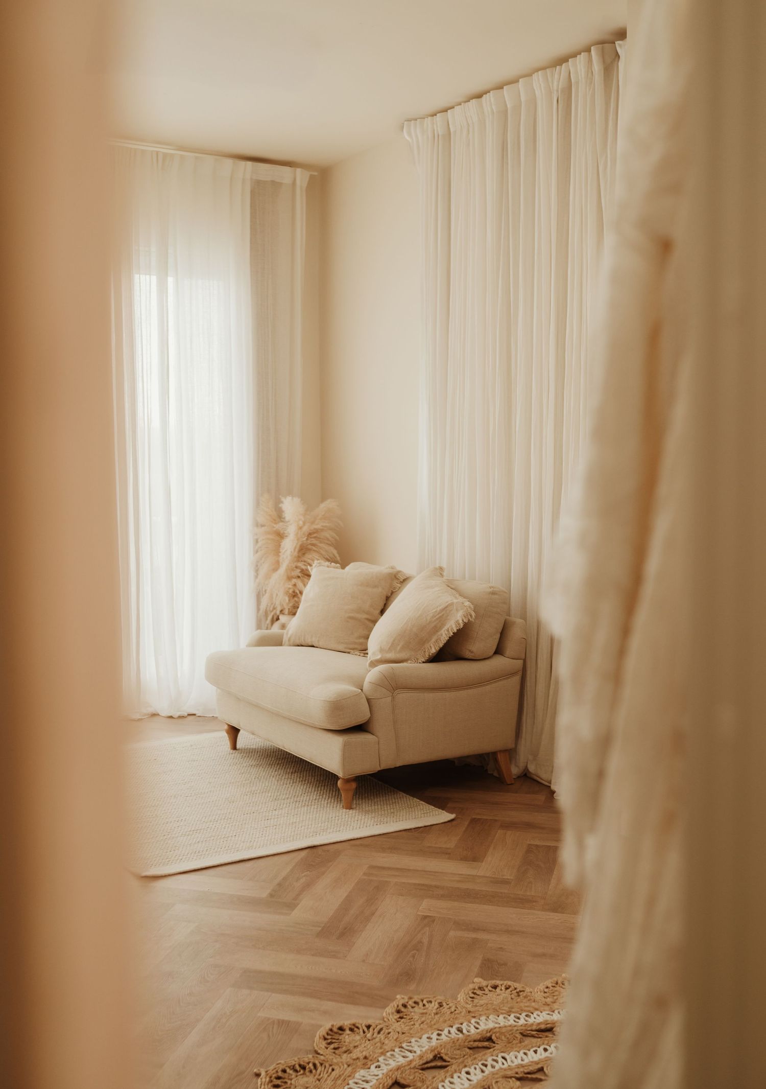 Light and airy interior with beige couch against white curtains and herringbone wood flooring in minimalist living space.