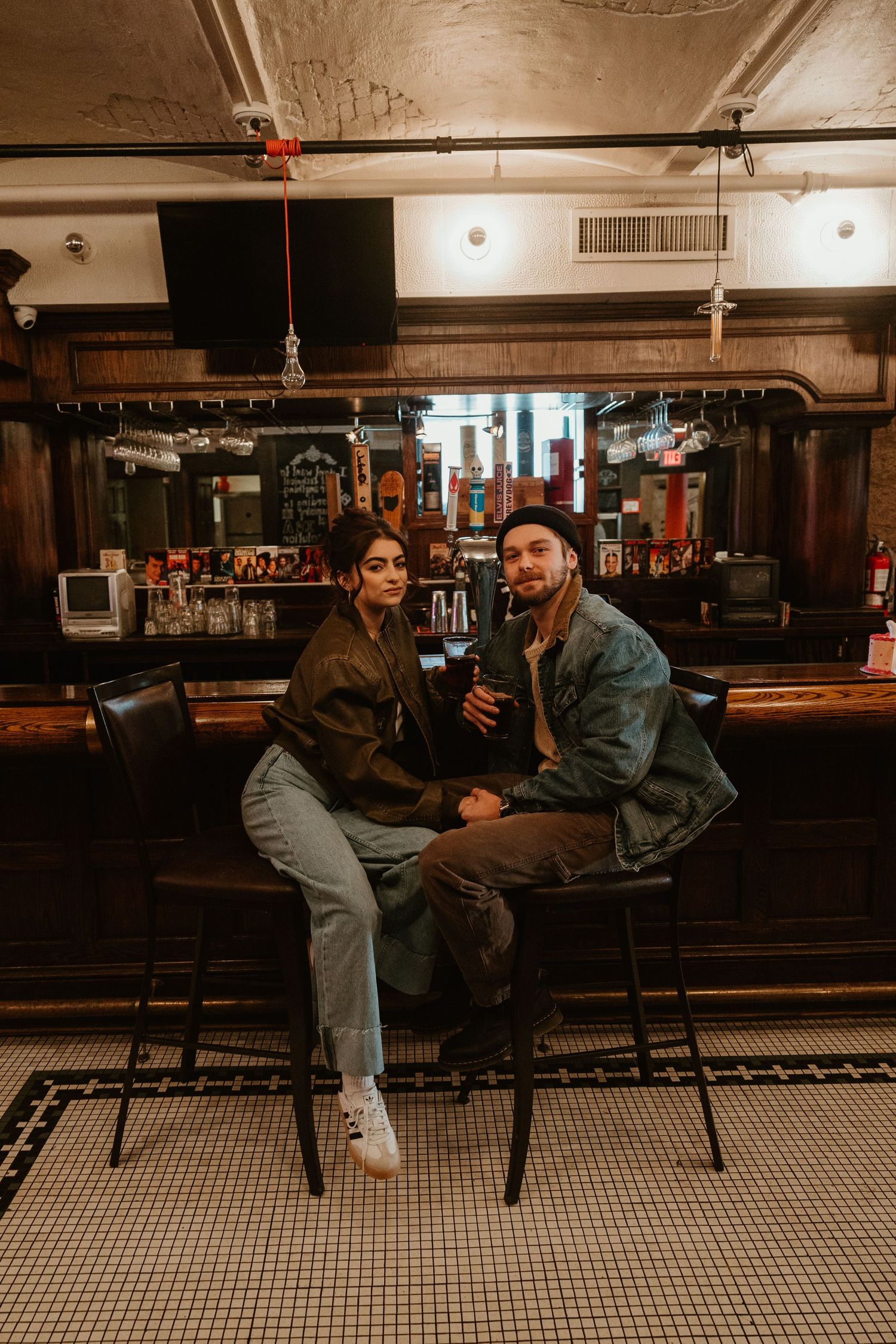 Two friends share a conversation while sitting at a vintage bar counter with black and white checkered floor tiles.