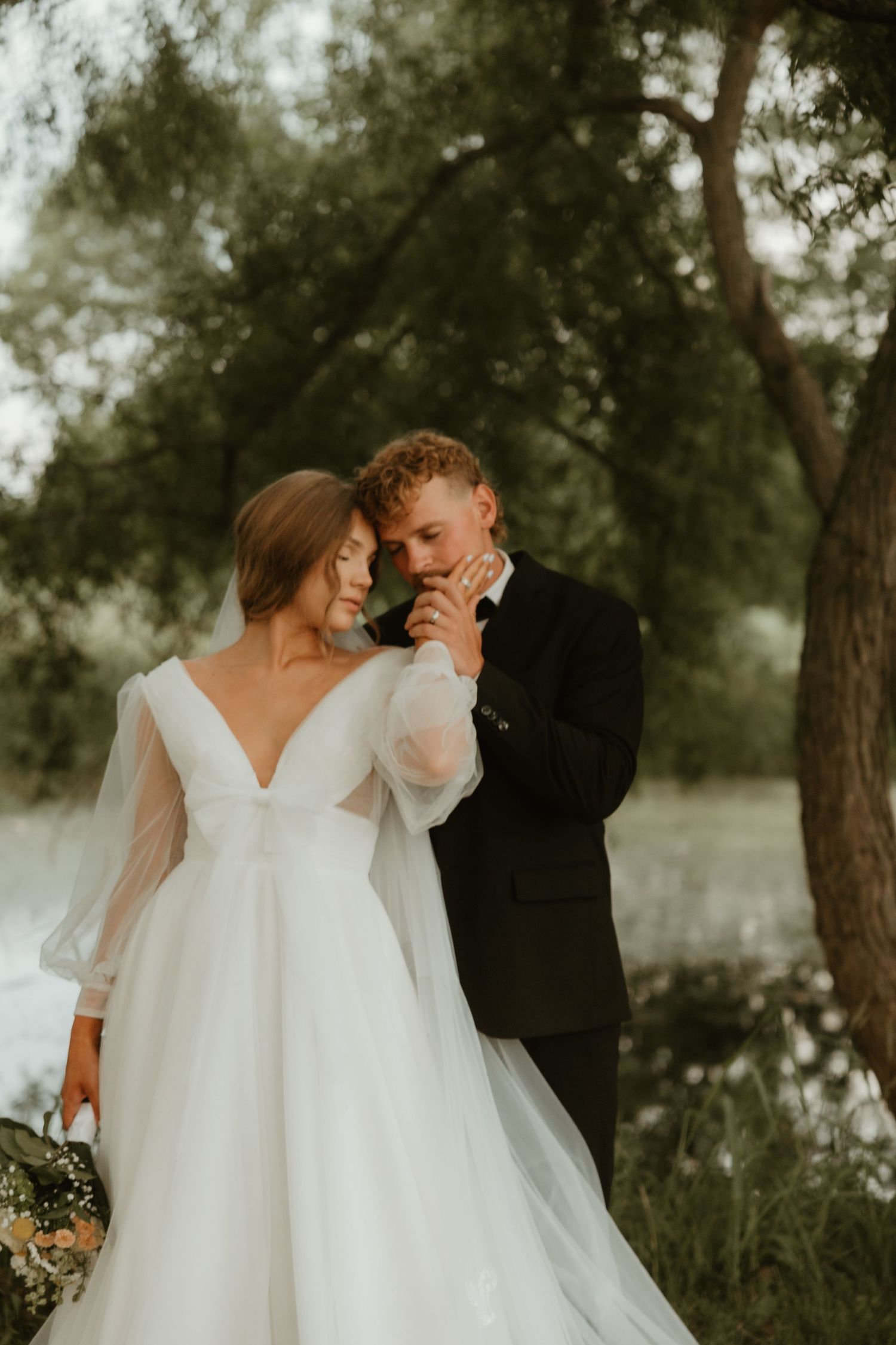 A romantic wedding couple shares intimate moments in a dreamy outdoor setting with trees and natural light.