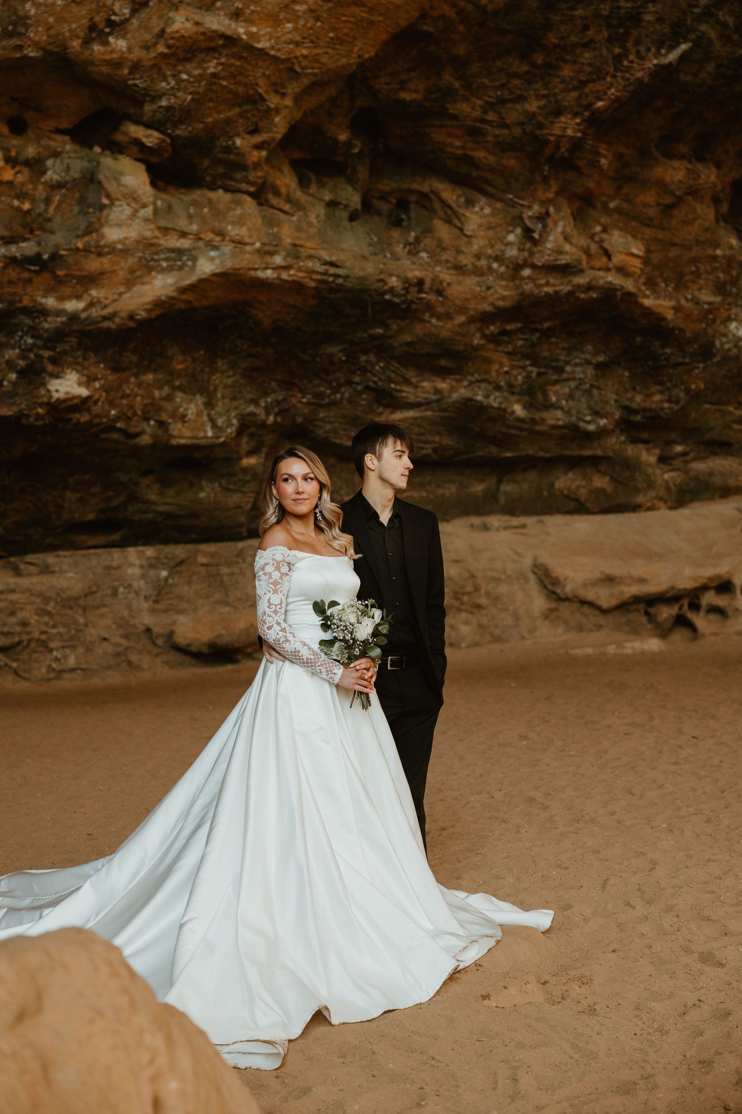 A couple in wedding attire poses together under a dramatic rocky cave overhang on a sandy beach.