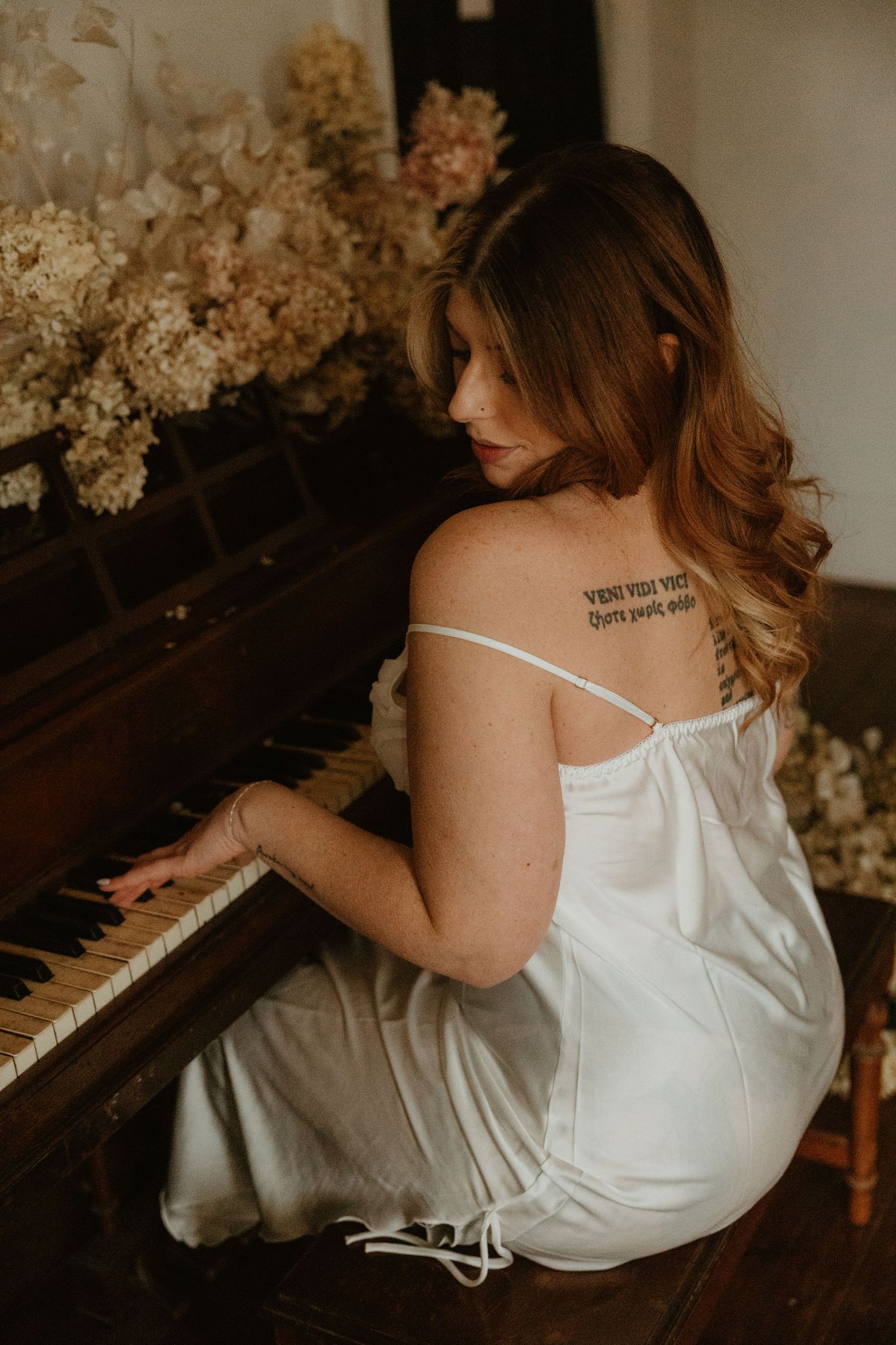Woman in white dress playing piano surrounded by dried flowers in moody lighting.