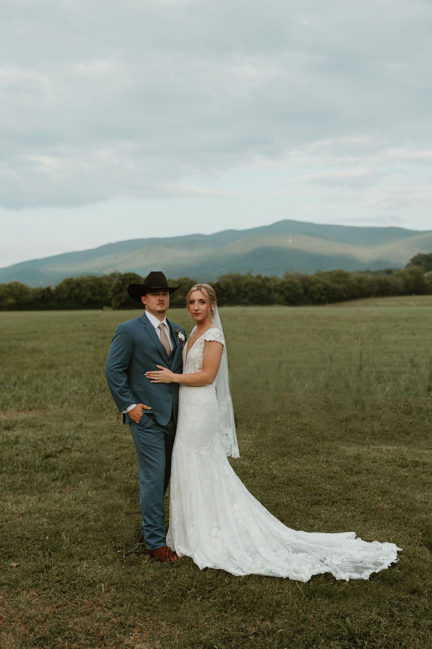 Newlyweds posing in an open field with mountain landscape backdrop.