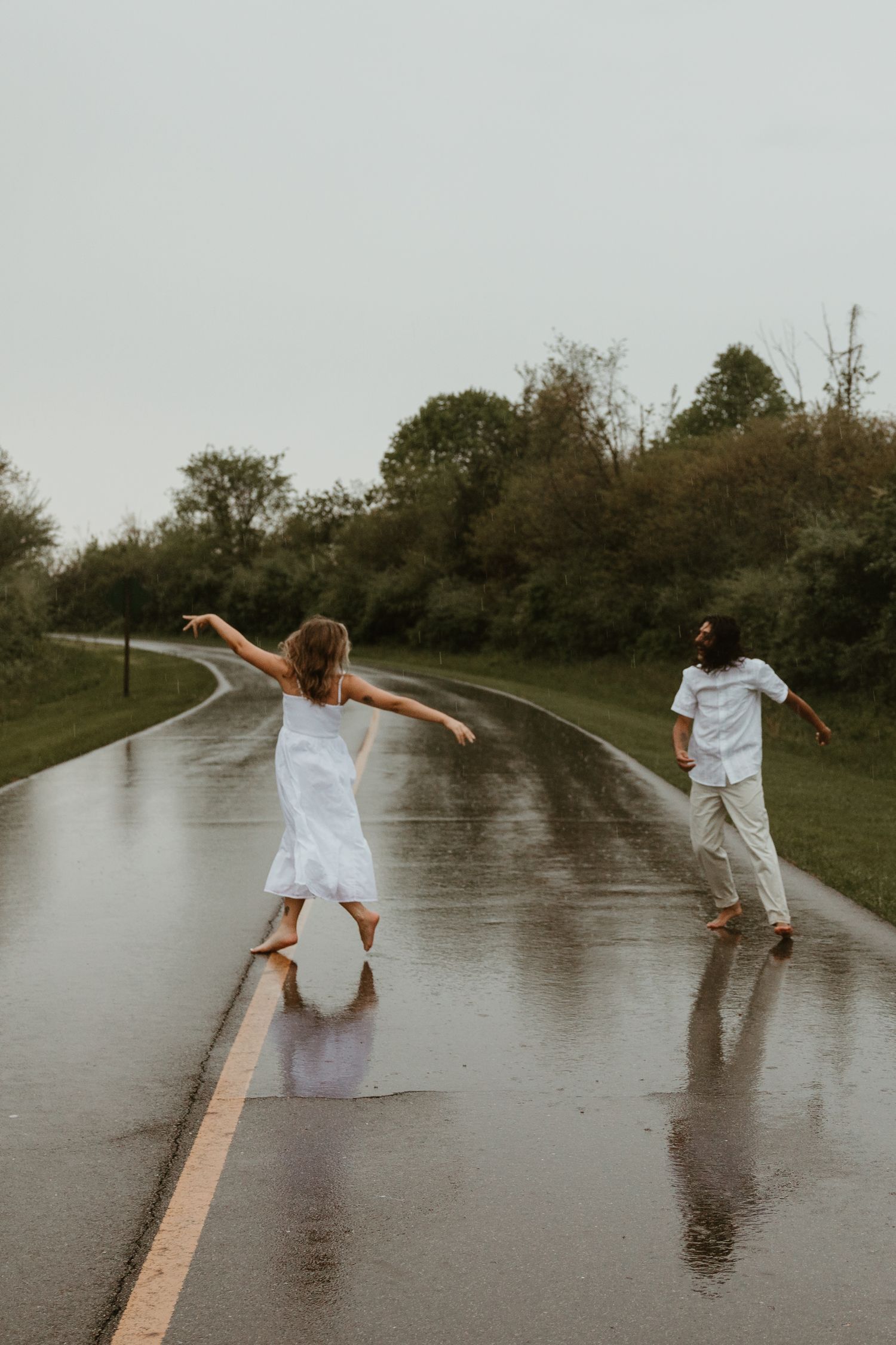 Two people in white outfits dance playfully in the rain on a wet road surrounded by green trees.