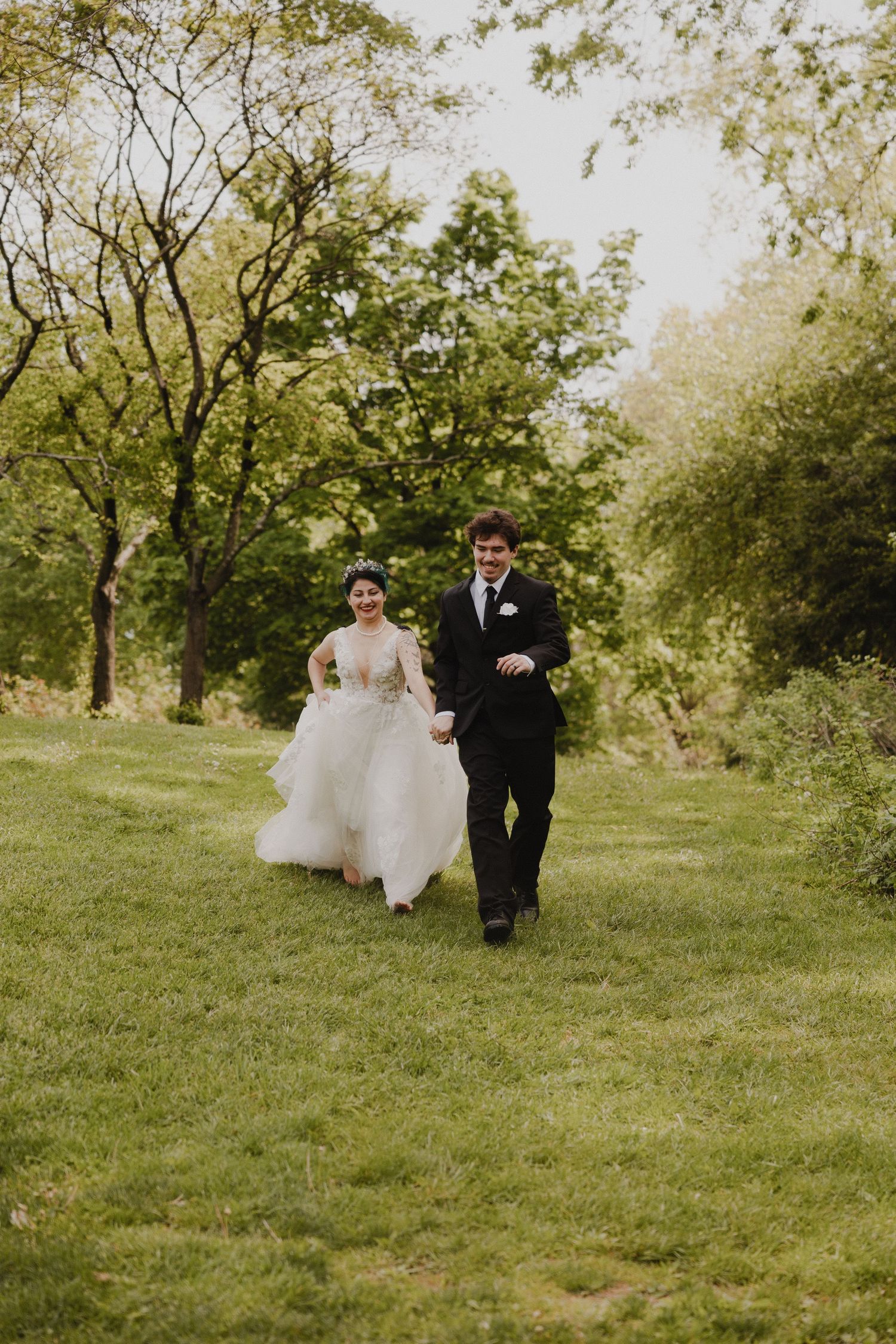 A couple in wedding attire walk together on a lush green lawn surrounded by trees on a sunny spring day.