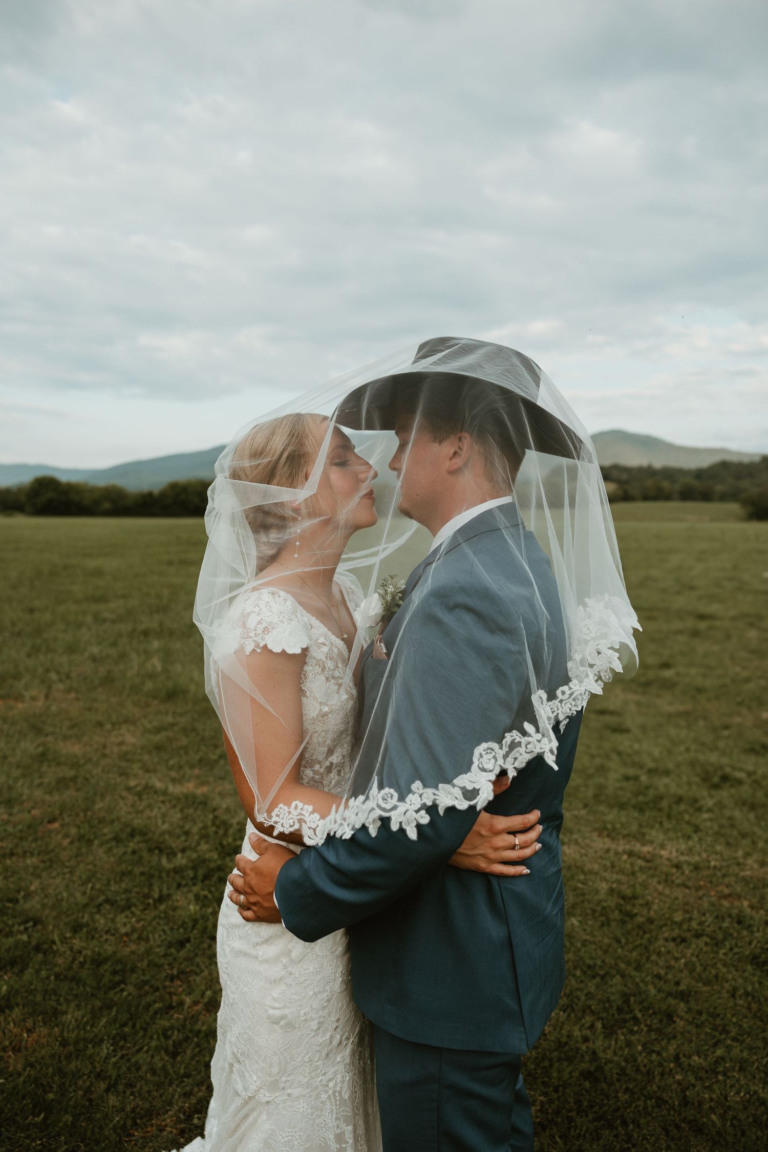 A romantic wedding couple embraces under a delicate lace veil against a scenic mountain landscape.