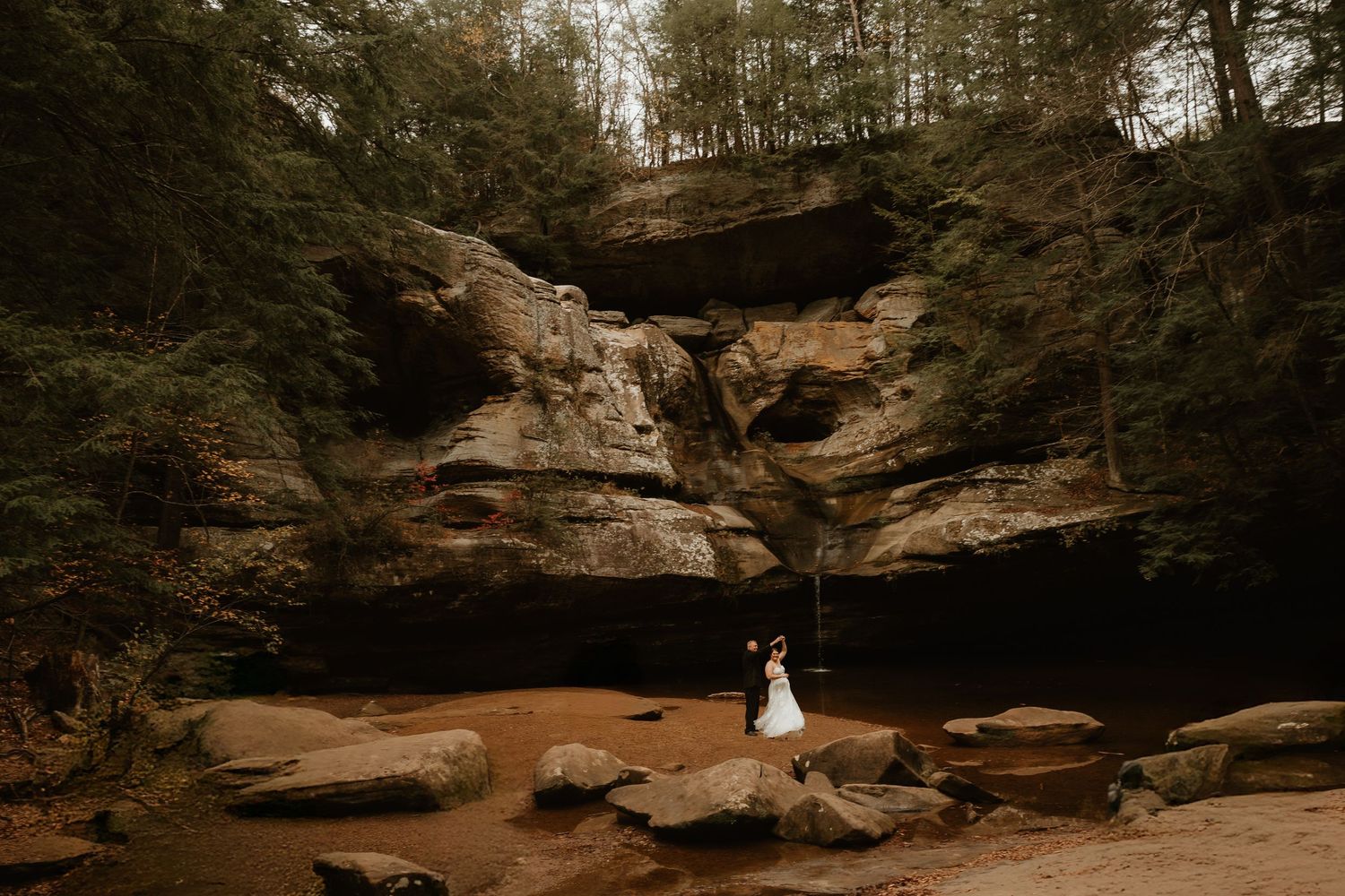 A couple in wedding attire stands beneath towering sandstone cliffs in a scenic natural canyon formation.