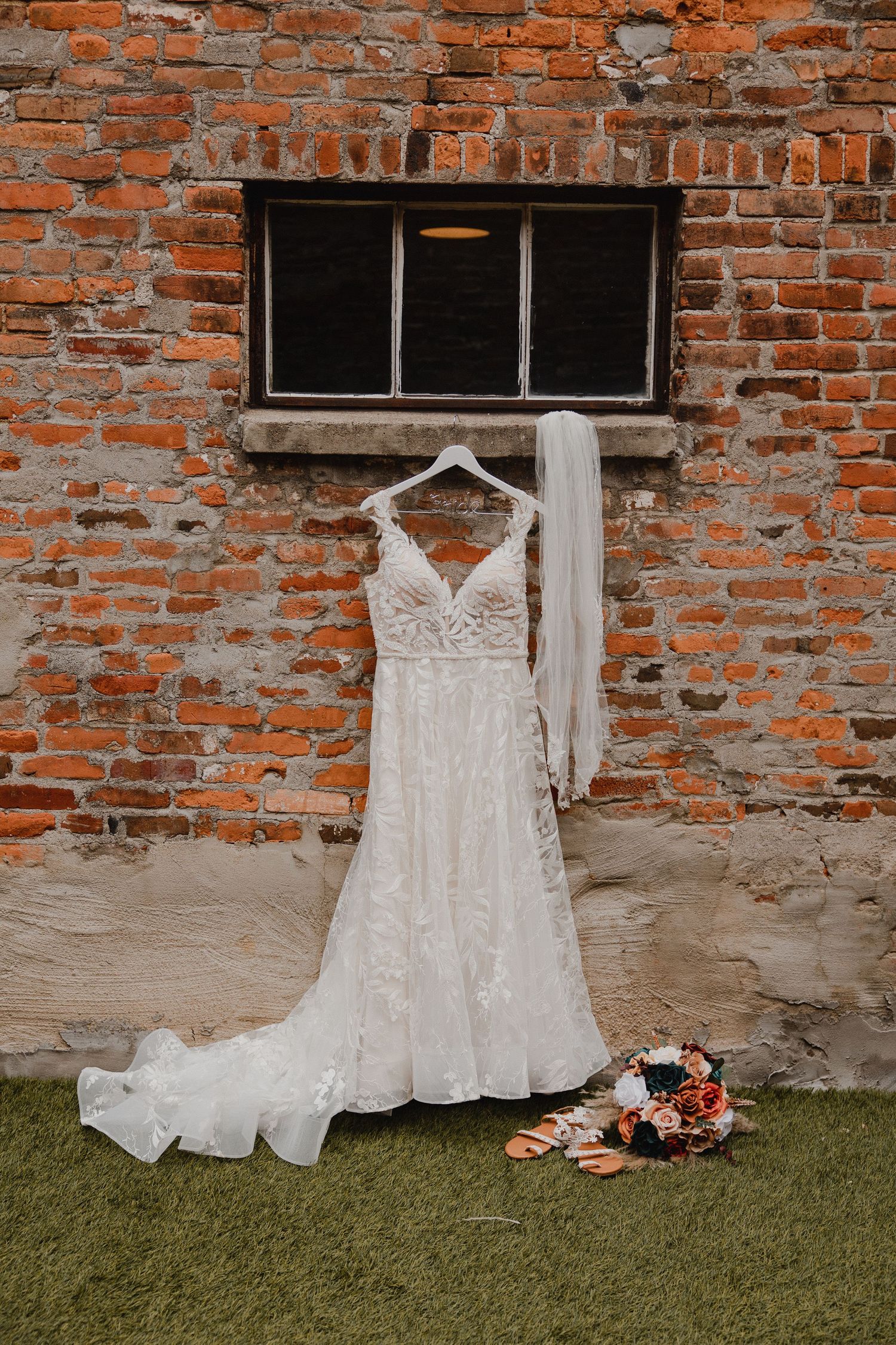 Elegant white lace wedding dress hanging on brick wall with floral bouquet on grass below.
