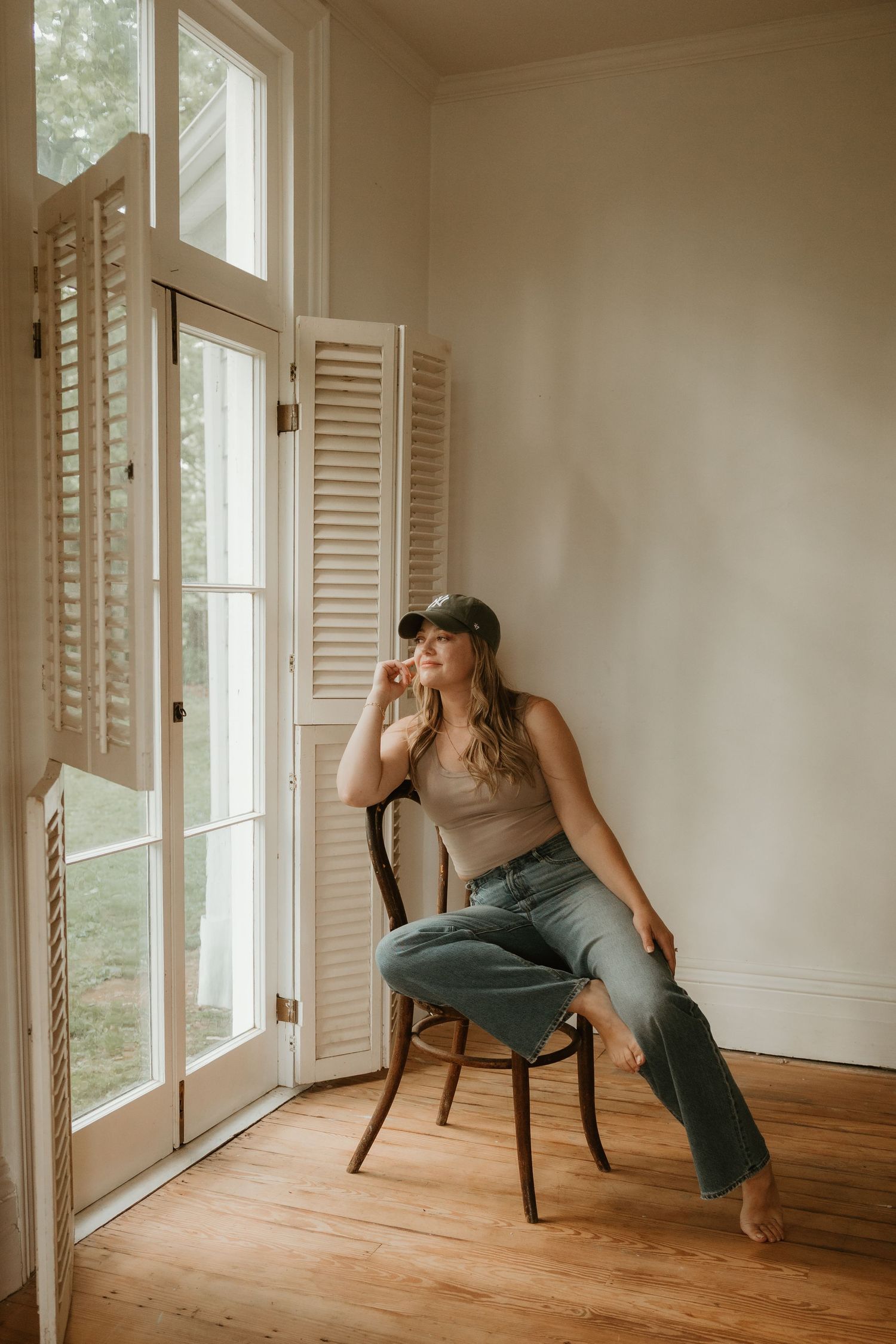 Person in casual attire sitting on wooden chair by bright windows with white shutters in minimalist room.