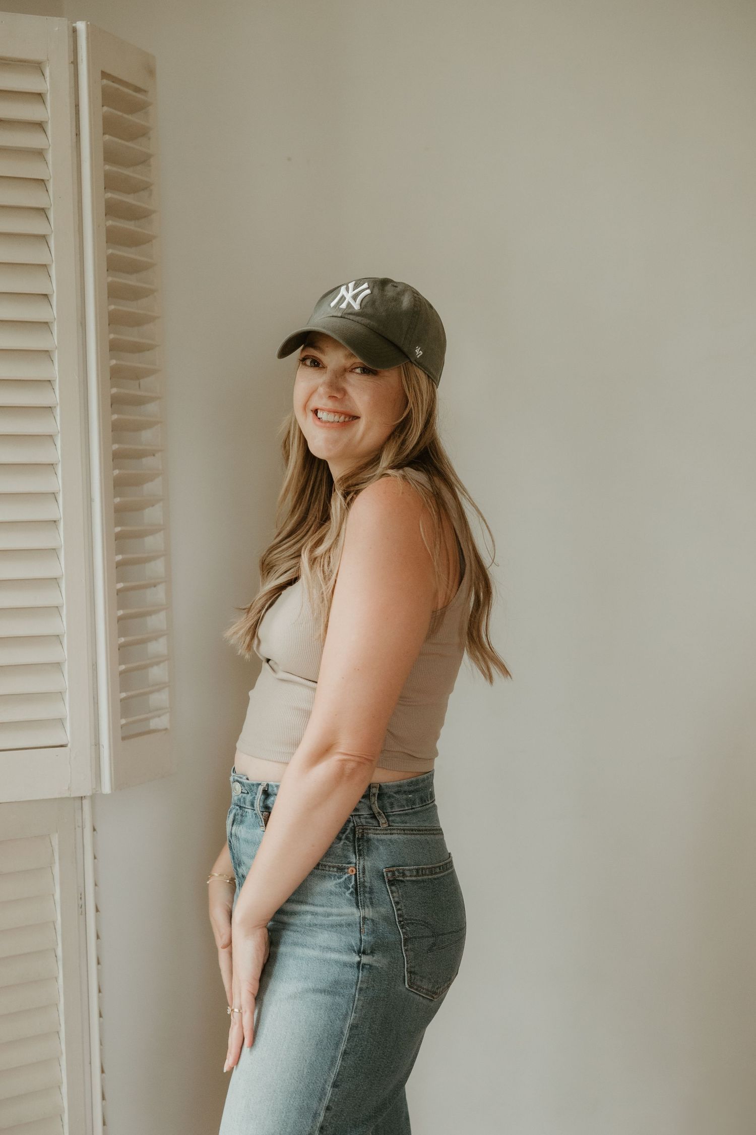 Someone wearing a Yankees baseball cap and denim jeans poses against a white wall with window shutters.