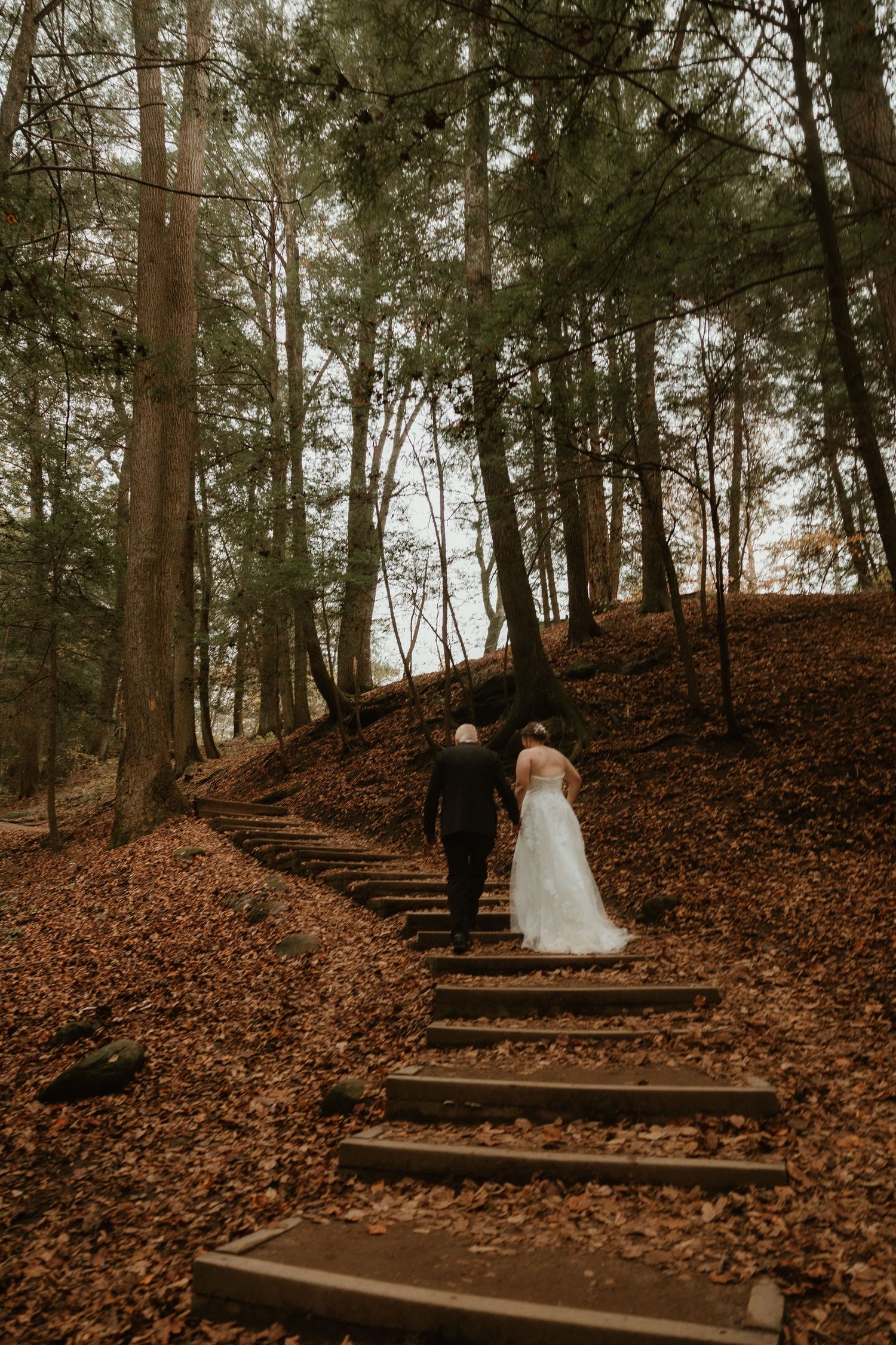 A couple walks up rustic wooden steps surrounded by fallen autumn leaves in a misty forest setting.