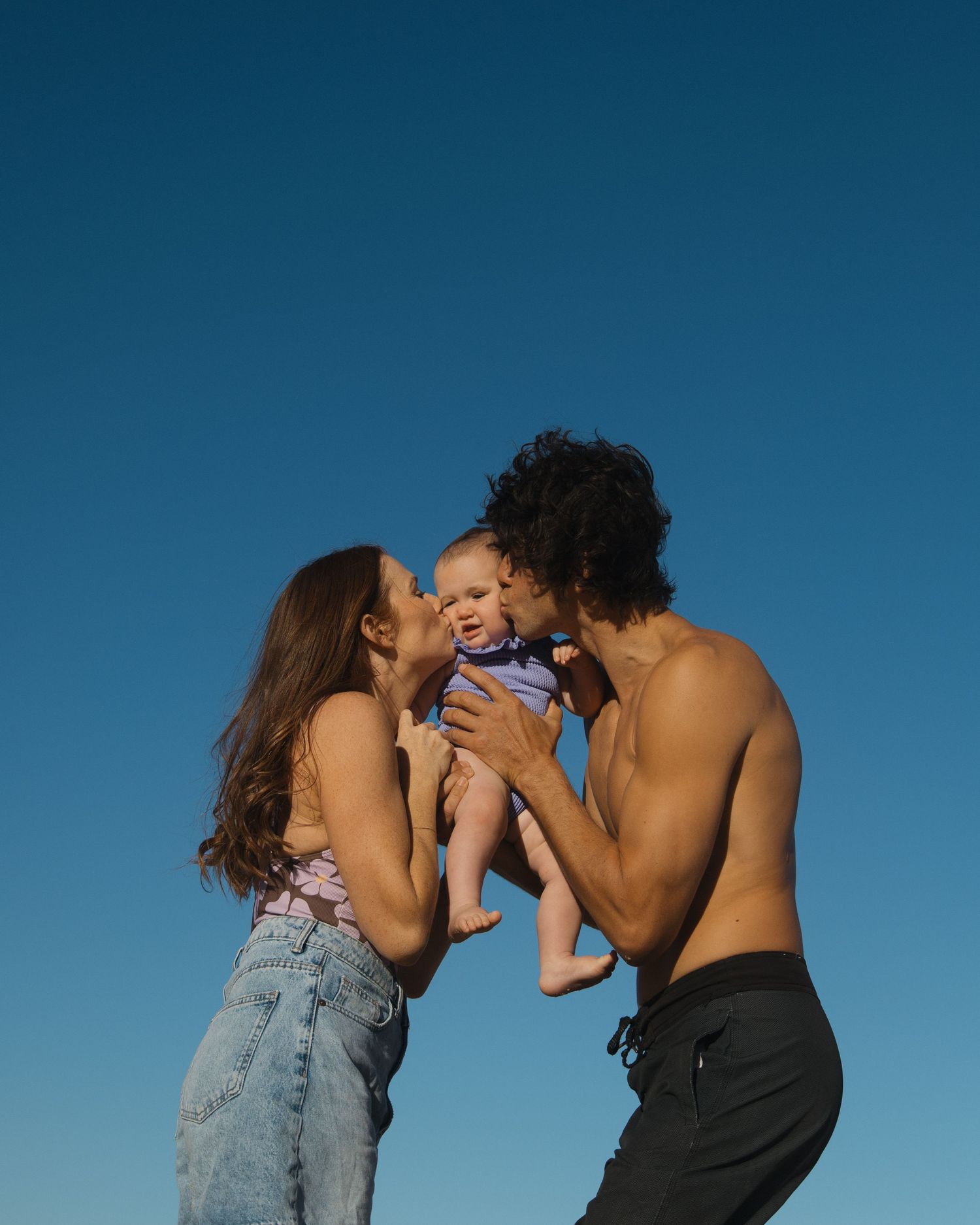 A couple kisses their smiling baby outdoors against a clear blue sky on a sunny day.