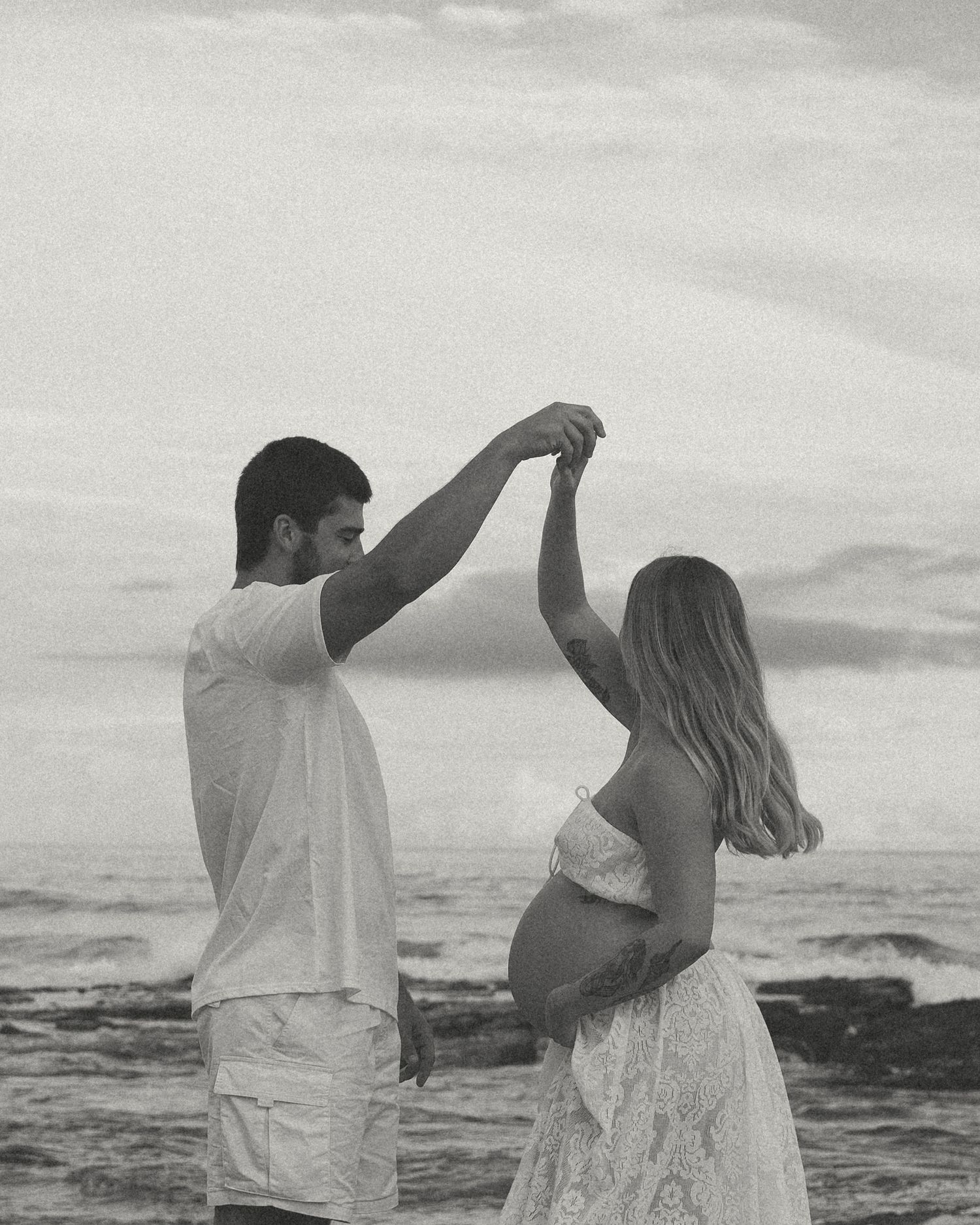 A couple in white outfits stands together on a beach at sunset, raising their arms in a celebratory pose.
