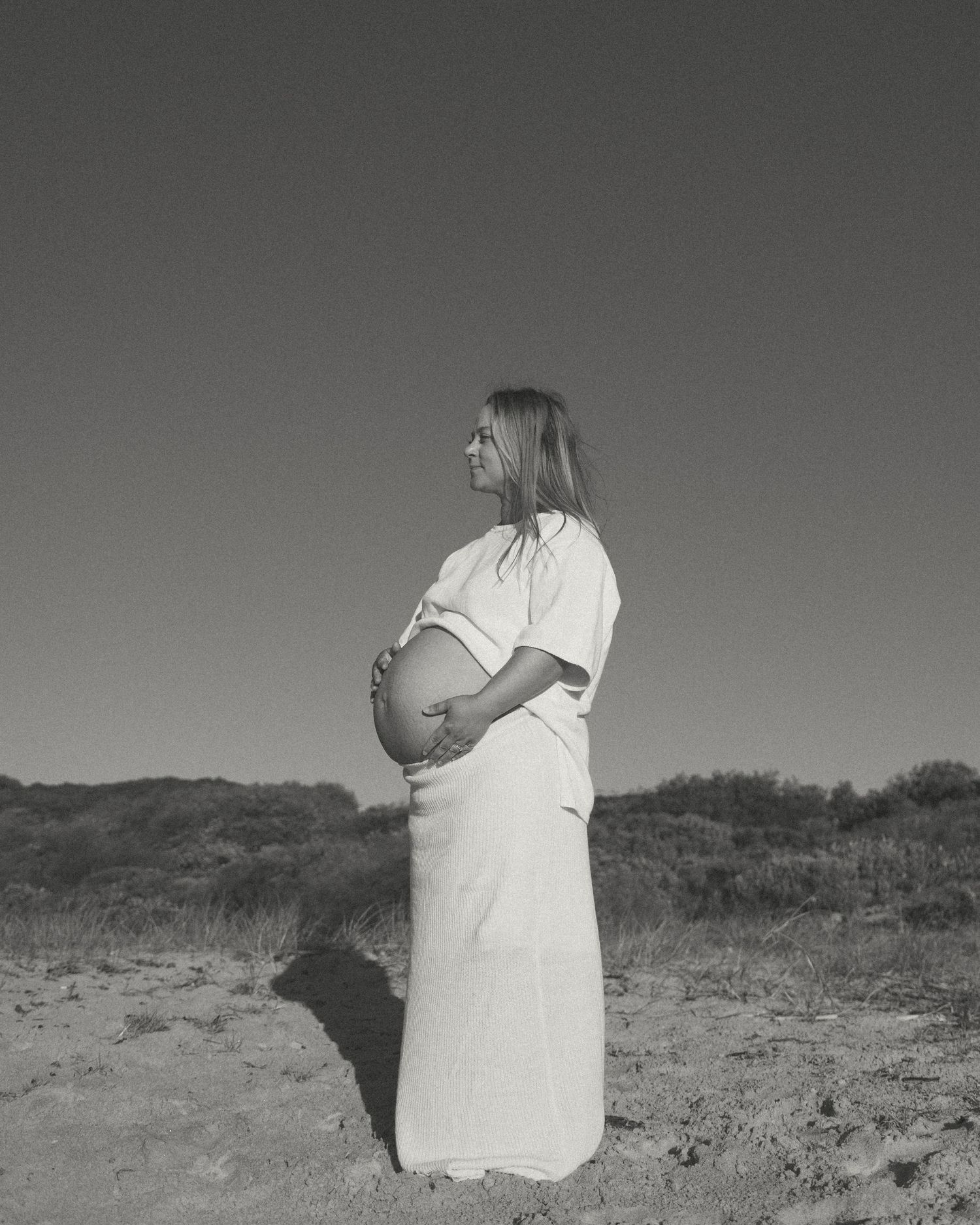 Black and white photo of a pregnant woman in a white dress cradling her belly outdoors in a natural landscape.