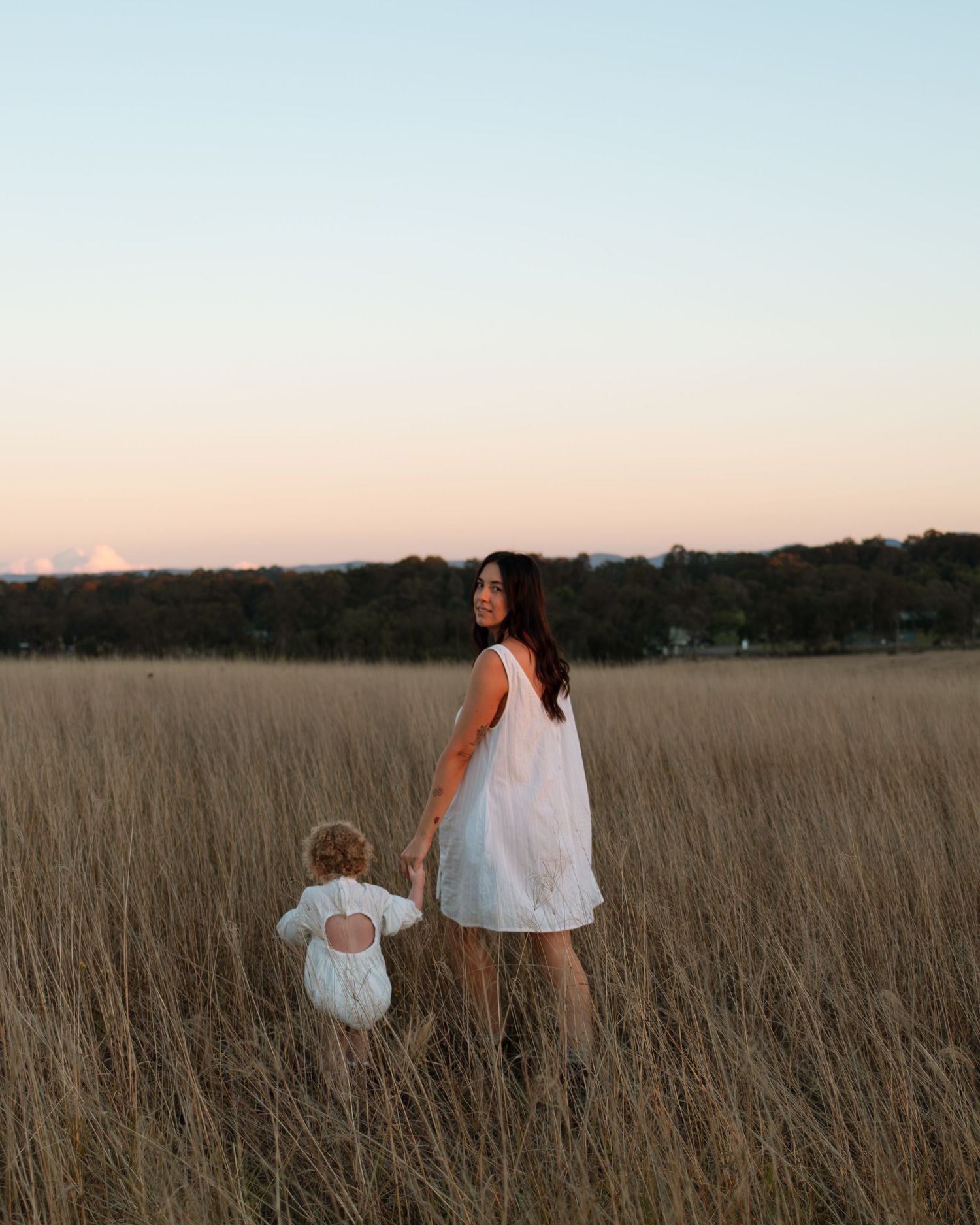 A woman in a white dress holds a toddler's hand in a golden field at sunset.