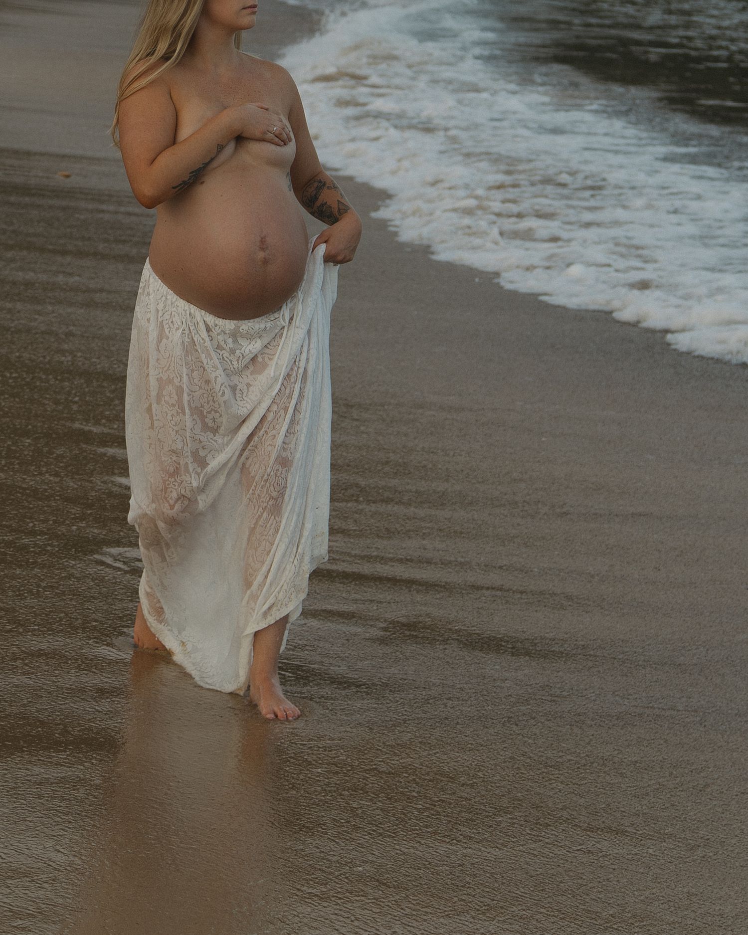 A pregnant figure in a flowing white dress stands at the edge of the ocean waves on a sandy beach at sunset.