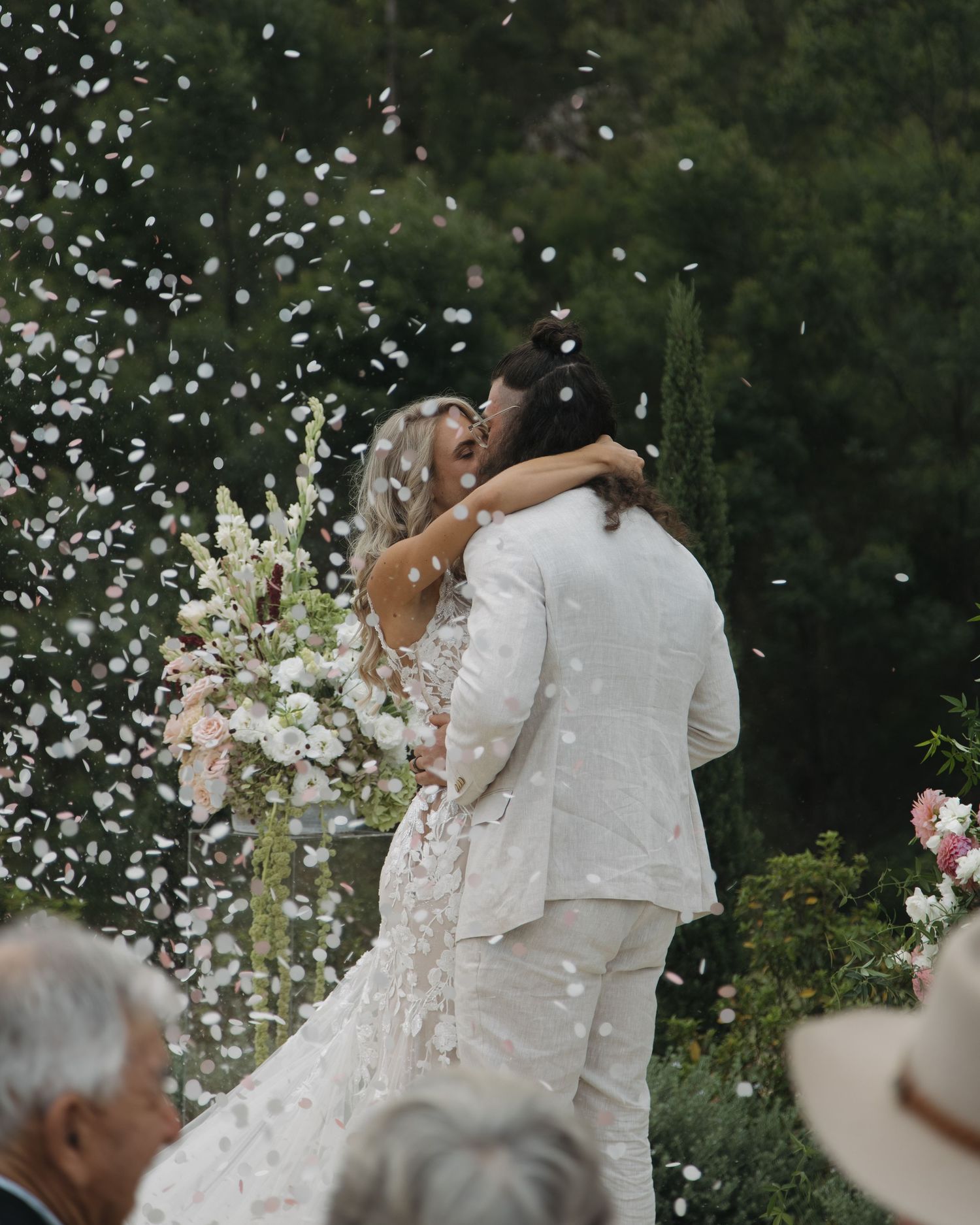 Newlywed couple sharing first kiss surrounded by white confetti petals at outdoor garden wedding ceremony.