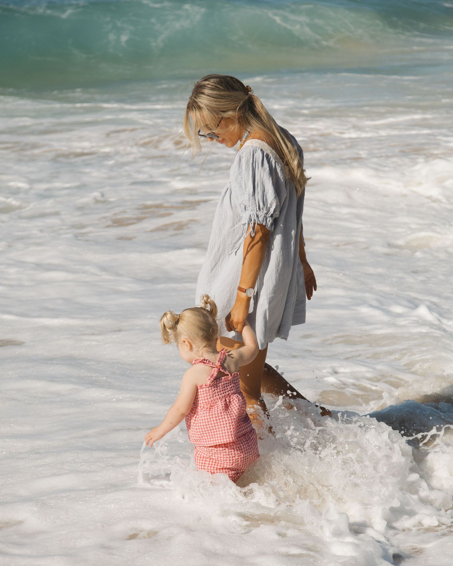 A blonde woman in a gray dress holds a toddler girl in a red gingham outfit while playing in ocean waves.