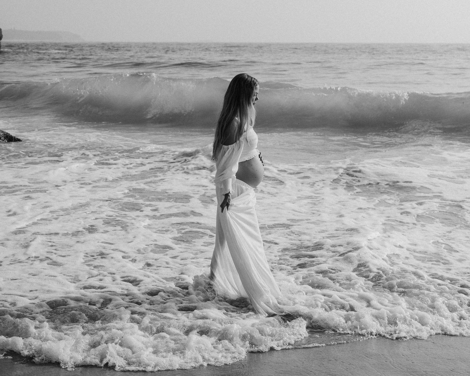 Person in white dress walking through ocean waves in black and white photography.
