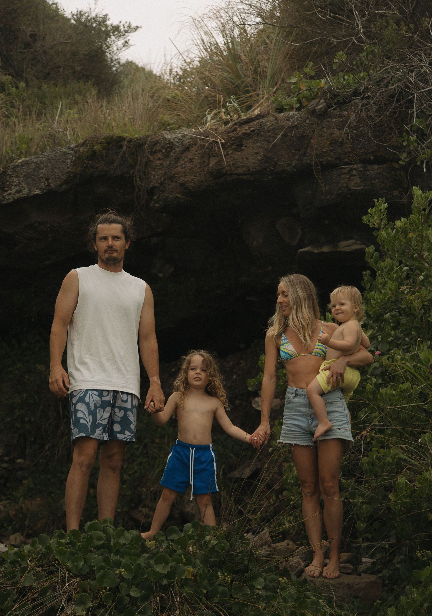 A family walks along a rocky cliff path surrounded by lush greenery on a cloudy day.